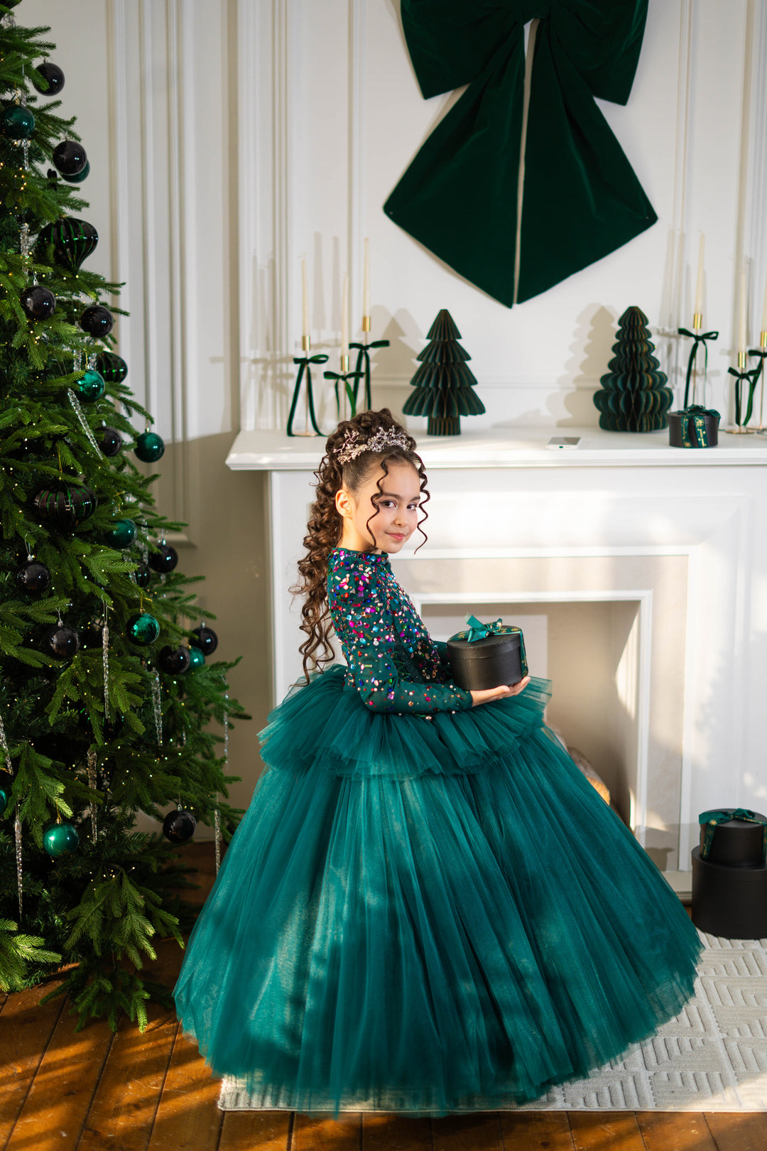 Young girl in a teal dress holding a small green tree in front of a decorated Christmas tree and fireplace.