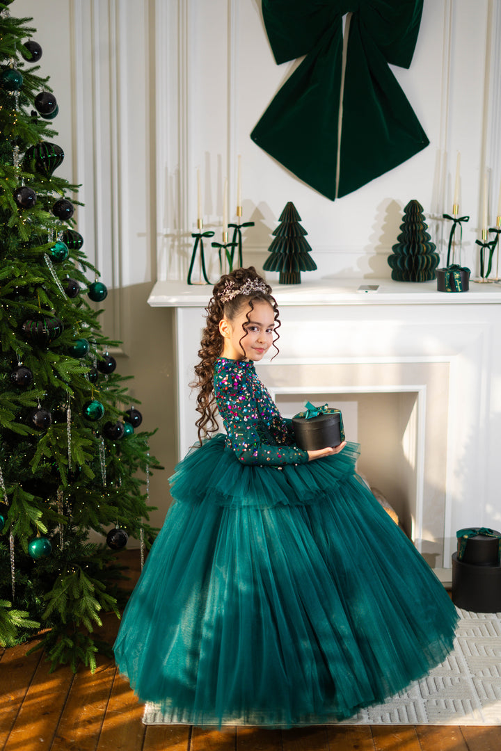 Young girl in a teal dress holding a small green tree in front of a decorated Christmas tree and fireplace.