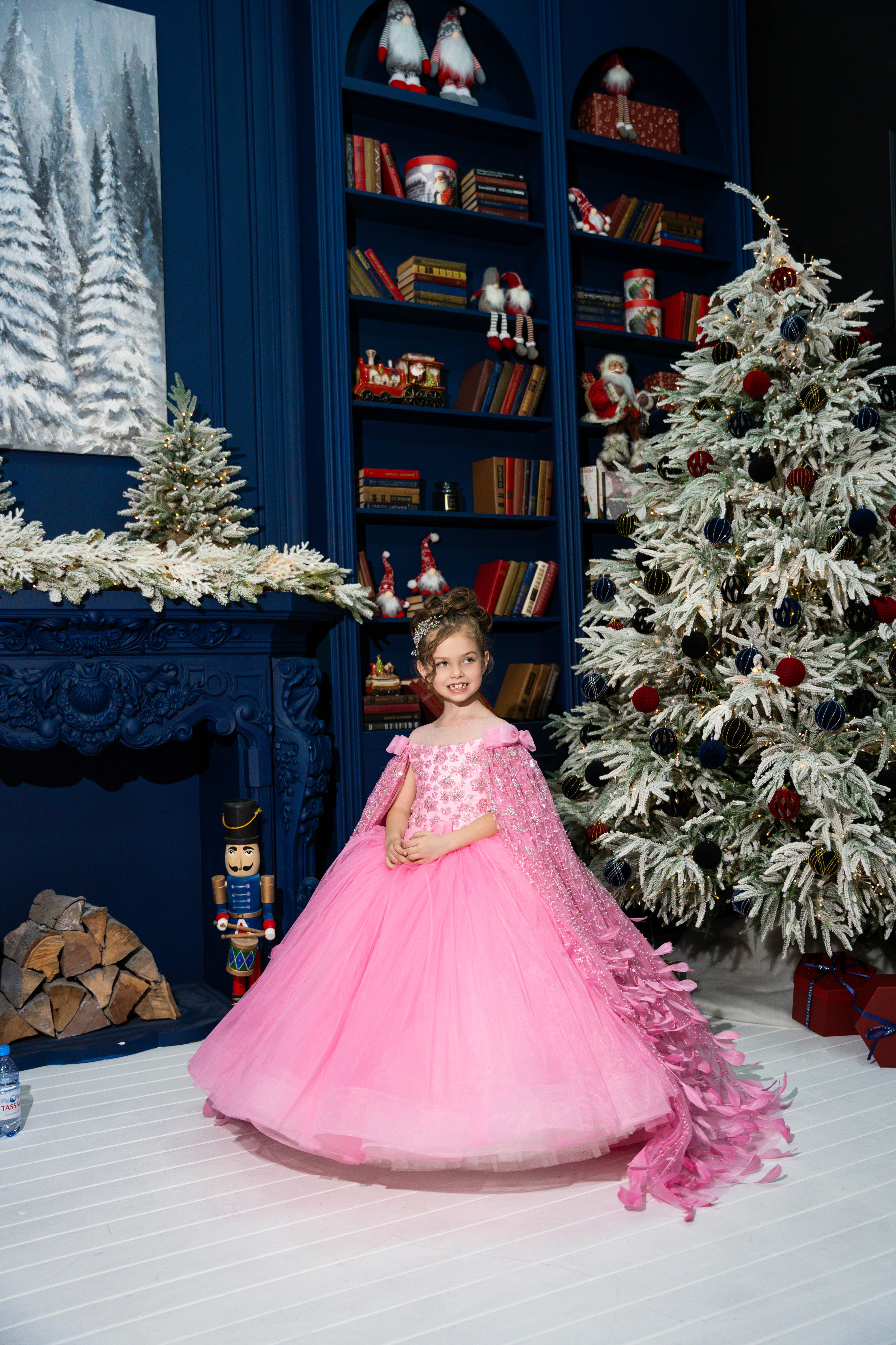Child in a pink dress standing in a festive room with a Christmas tree and decorated bookshelves.
