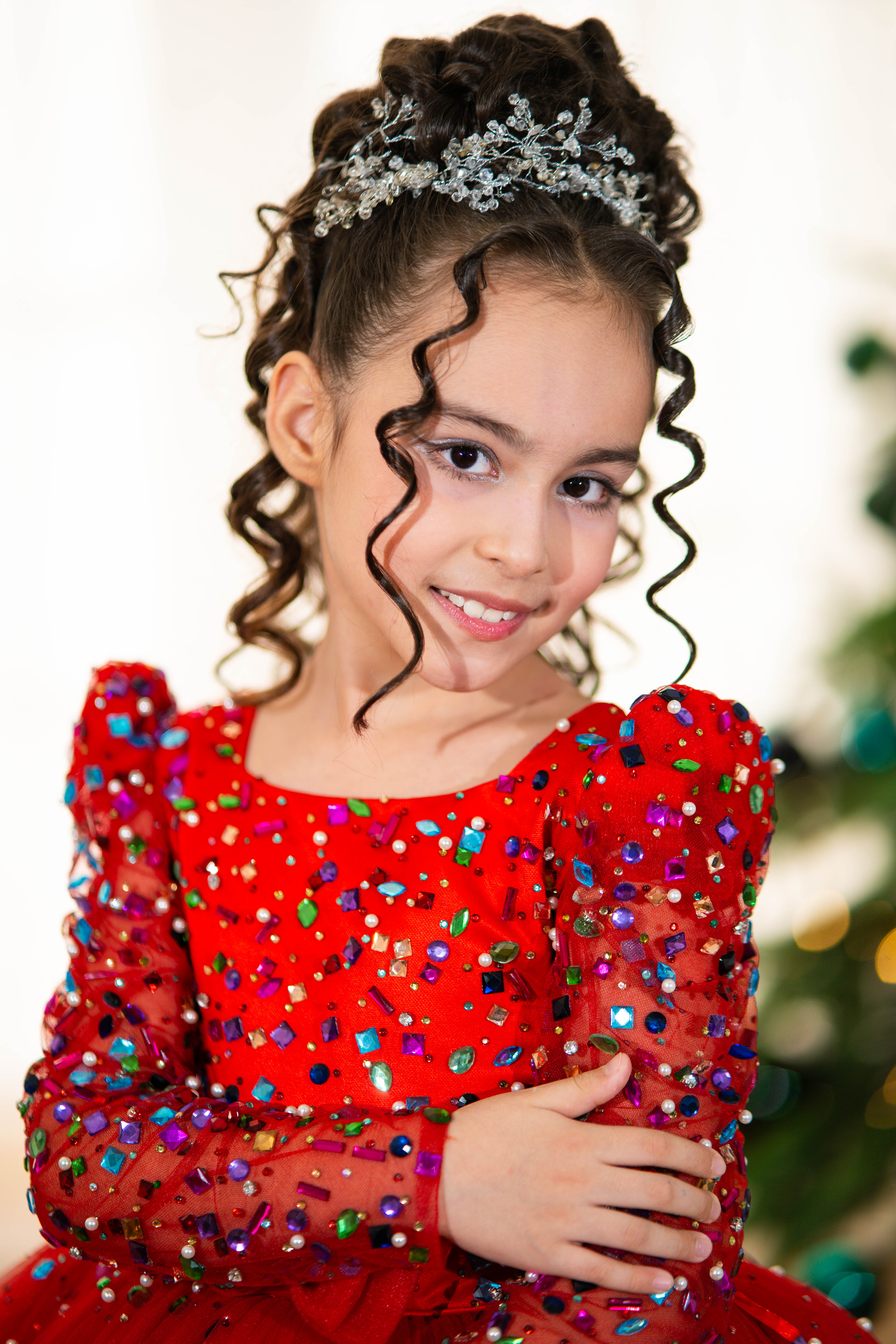 Young girl in a red dress with colorful sequins, wearing a silver headband, against a blurred background.