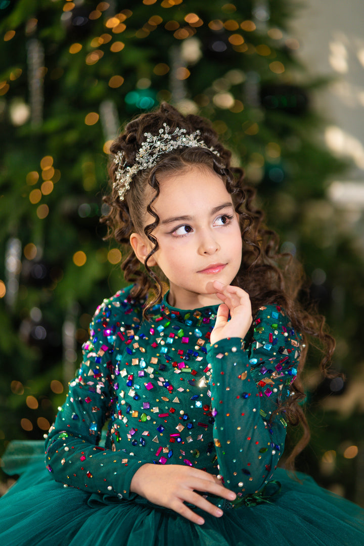 Young girl in a green sequined dress with a tiara, standing in front of a decorated Christmas tree.