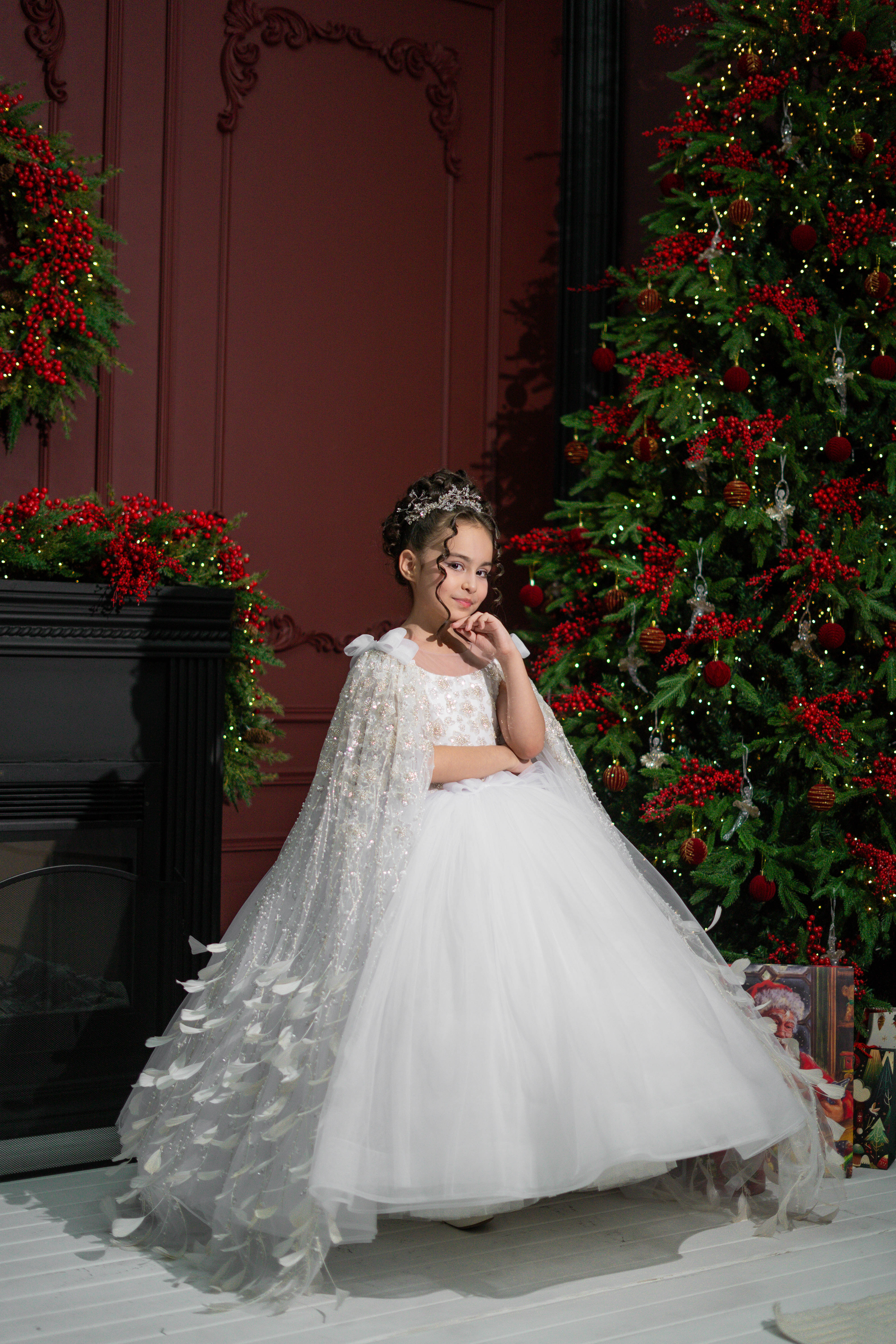  A child in a white dress with a cape standing in front of a decorated Christmas tree. 