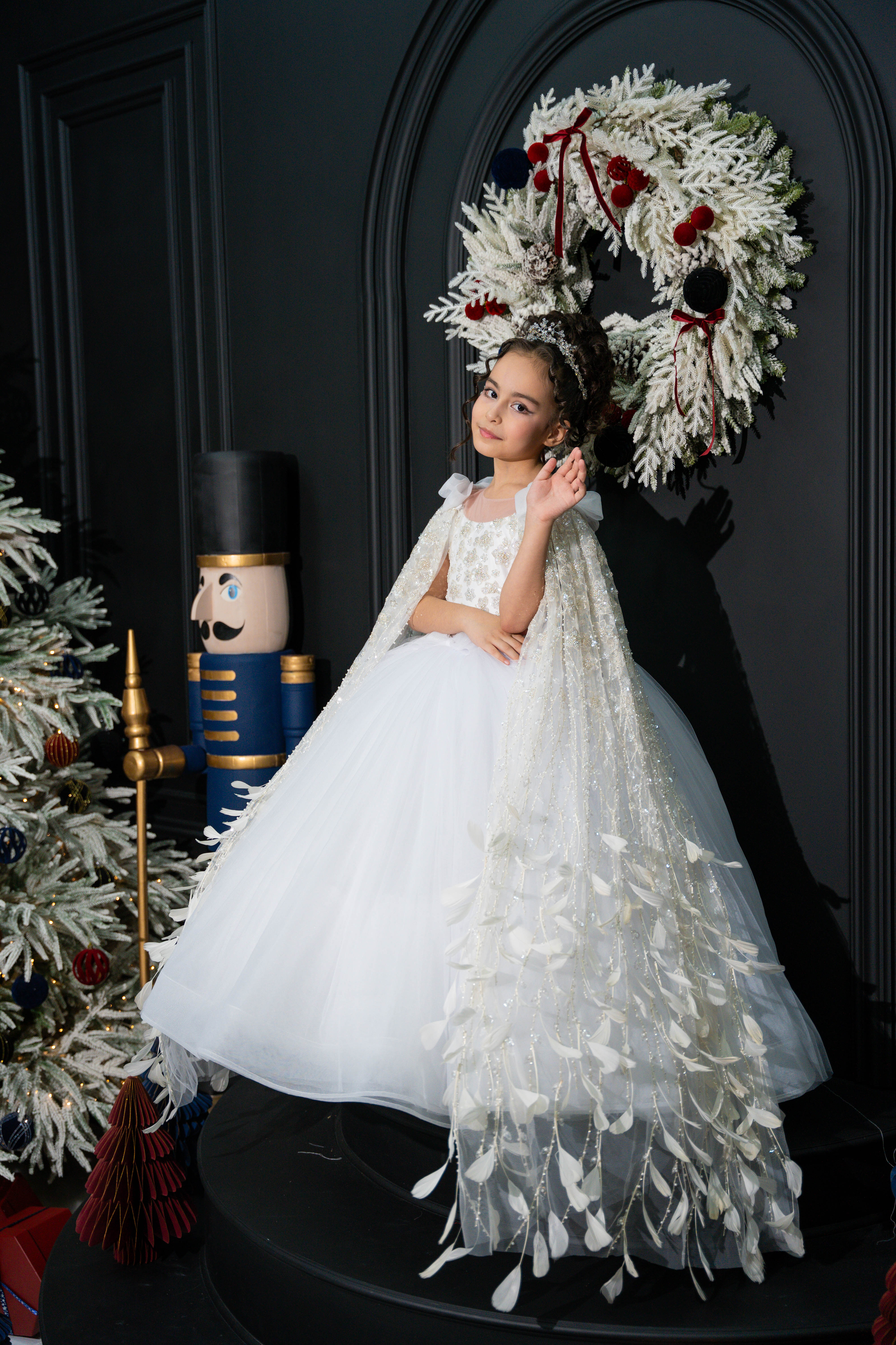 A child in a white dress with a decorative wreath and Christmas decorations in the background