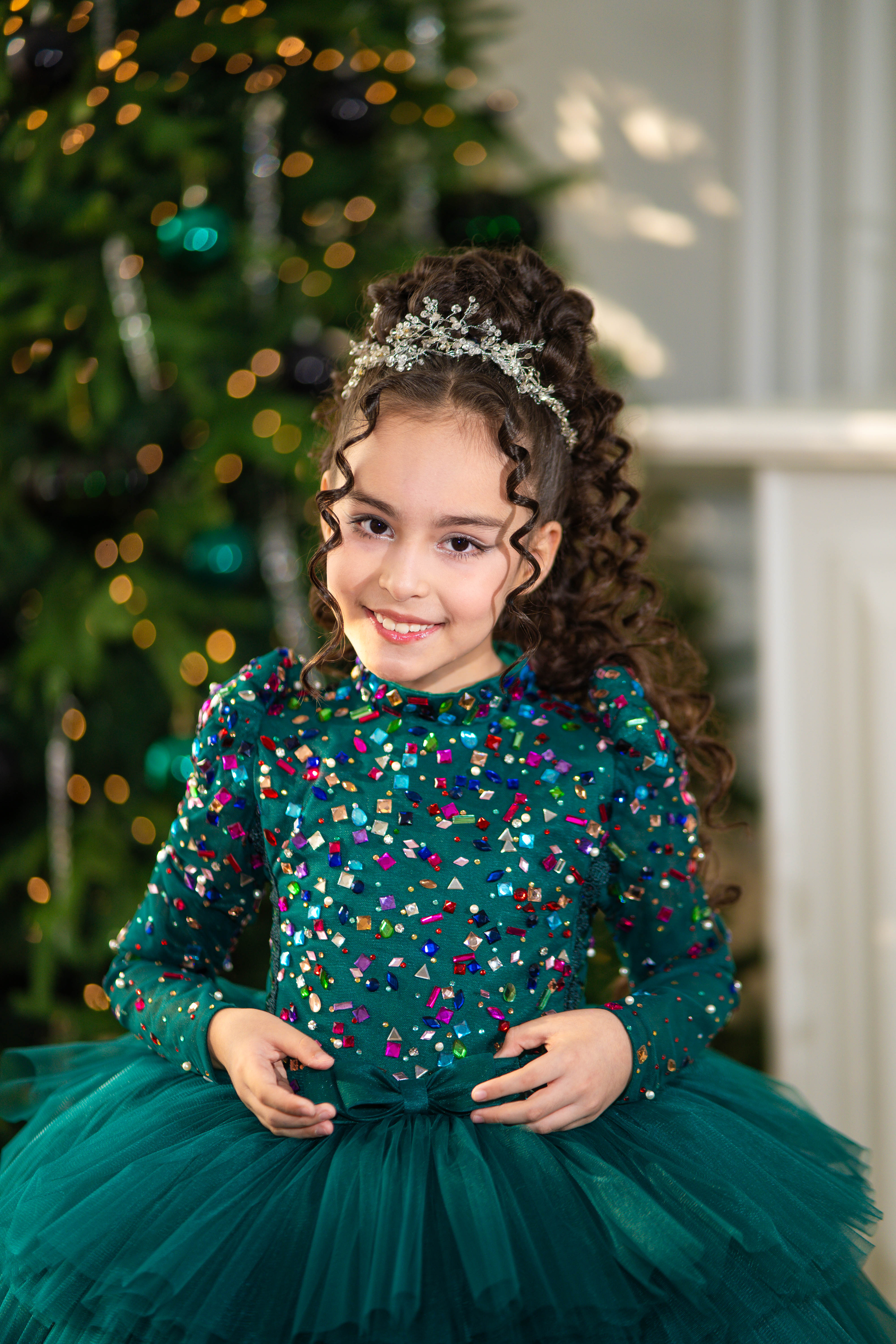 Young girl in a green dress with colorful sequins standing in front of a decorated Christmas tree.
