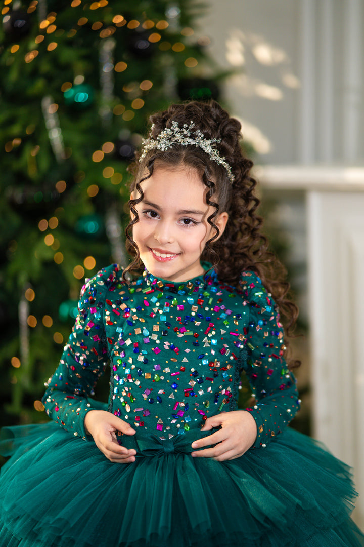 Young girl in a green dress with colorful sequins standing in front of a decorated Christmas tree.