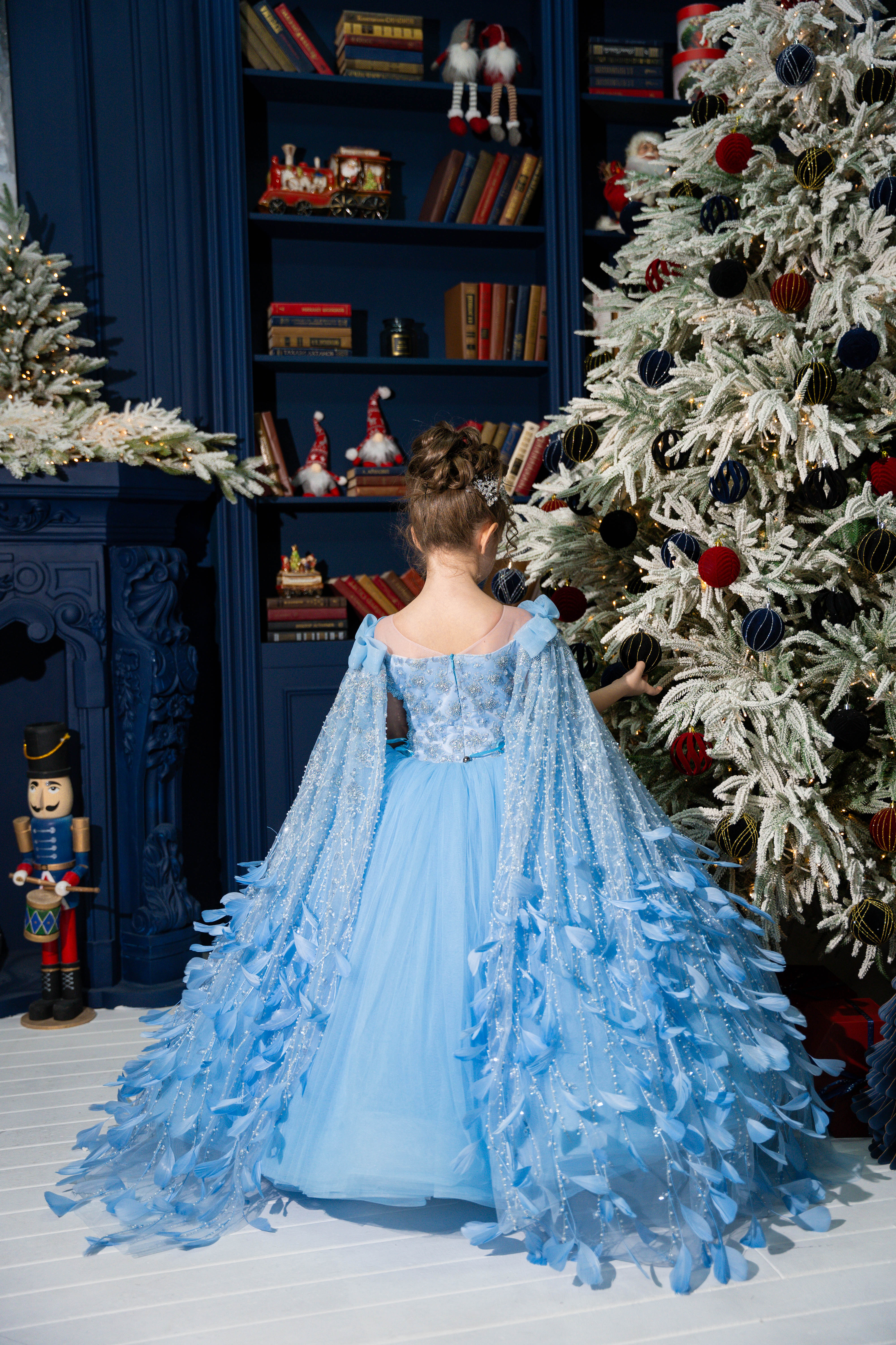A girl in a blue dress standing in front of a decorated Christmas tree and bookshelf.