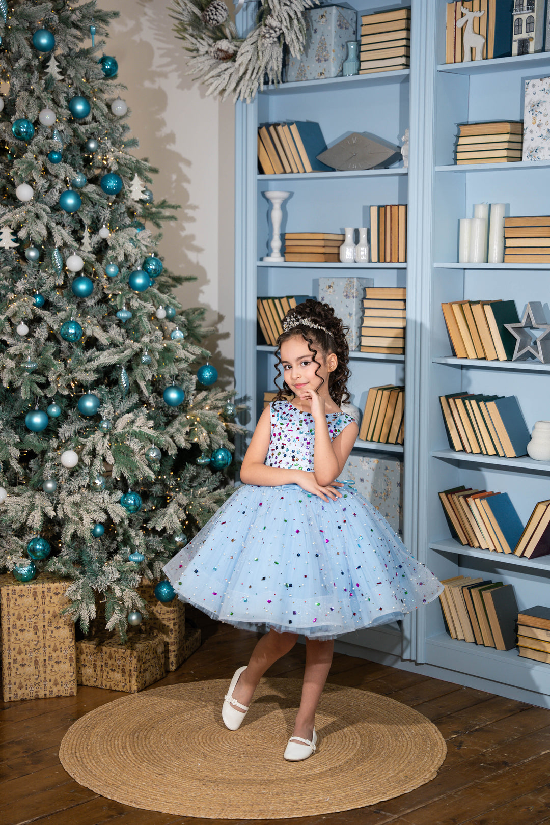 Young girl in a polka dot dress standing in front of a Christmas tree and bookshelf.