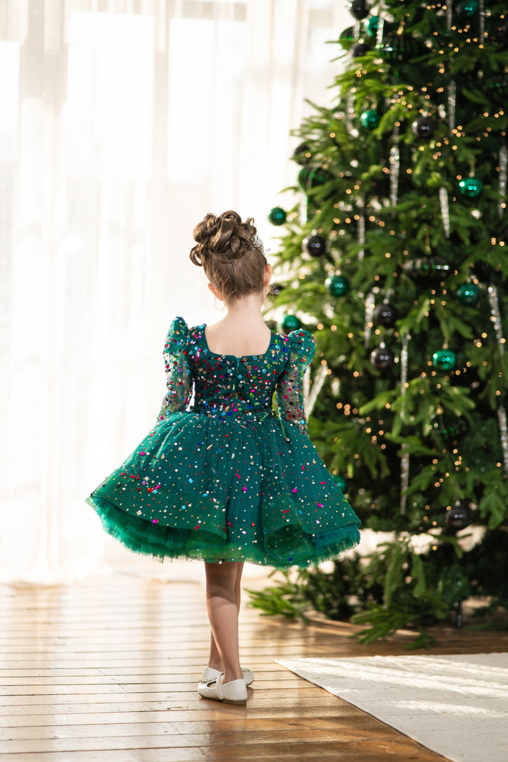 Child in a green dress standing in front of a decorated Christmas tree.