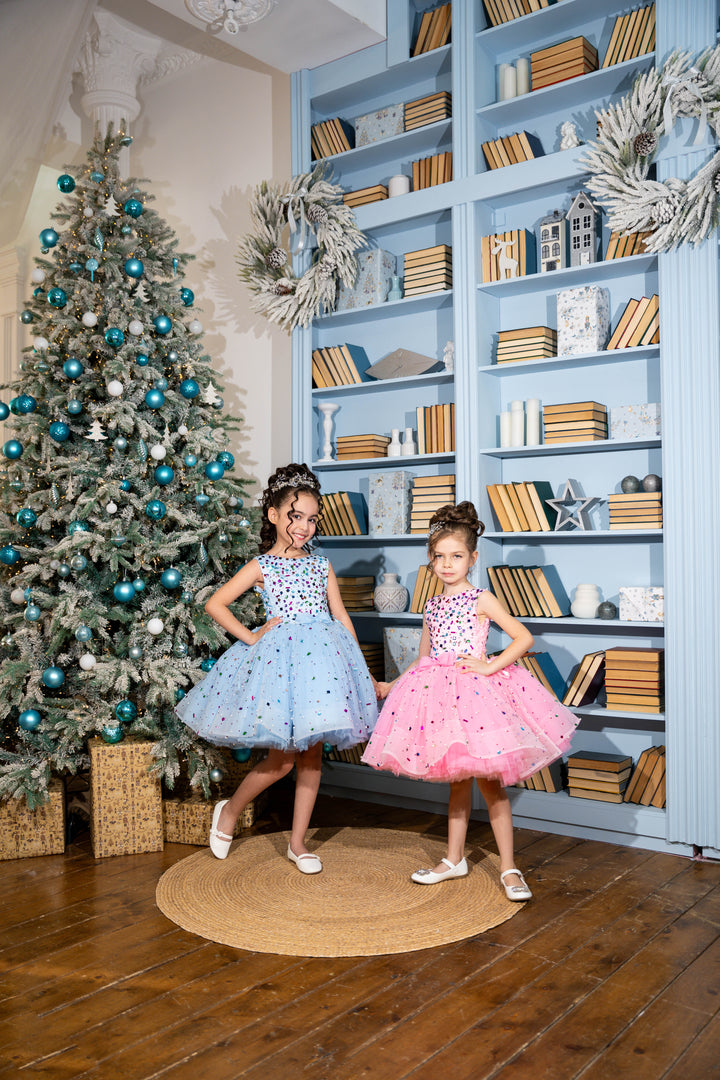 Two young girls in dresses standing in a room with a Christmas tree and bookshelf.