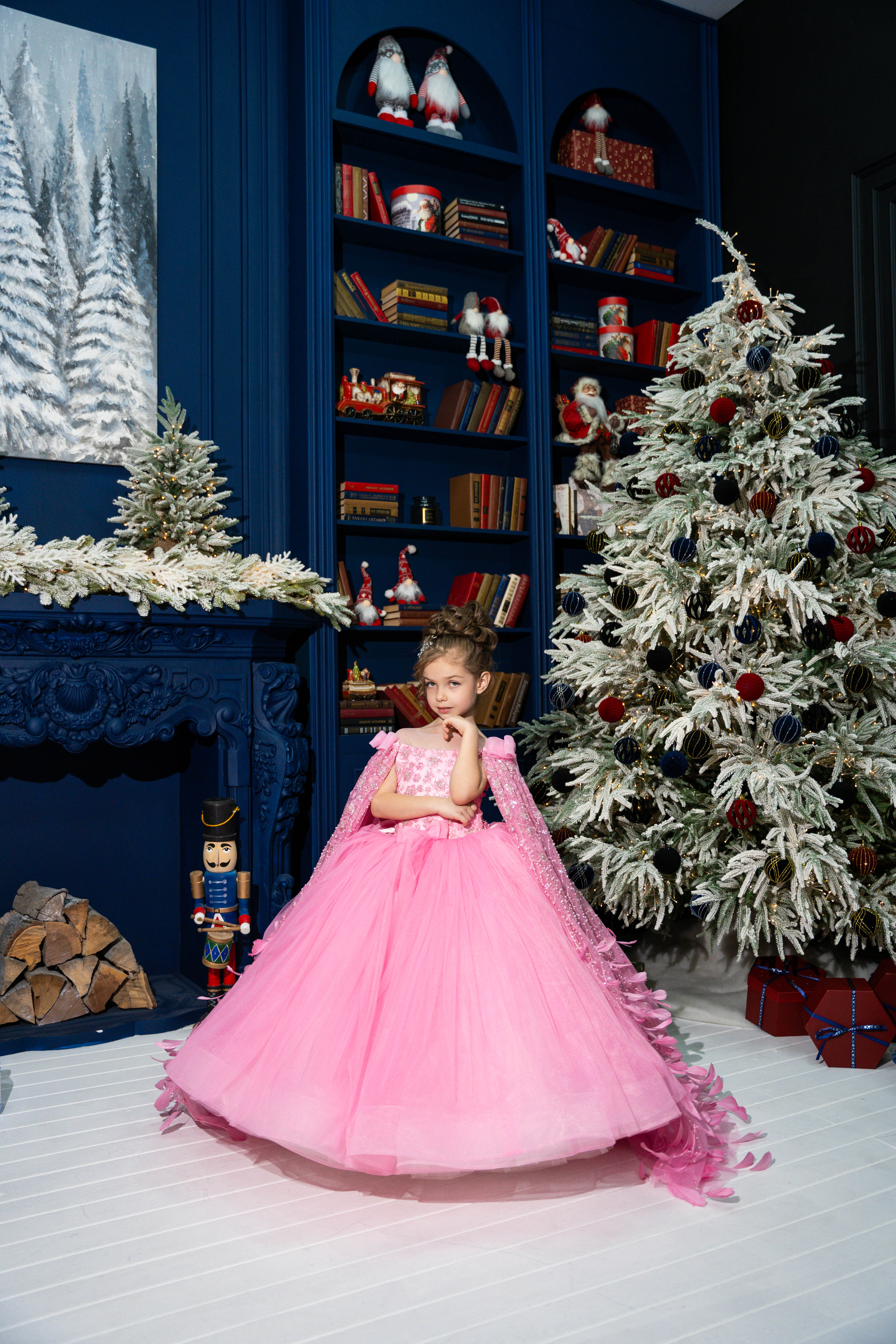 Child in a pink dress standing in a festively decorated room with a Christmas tree and bookshelf.