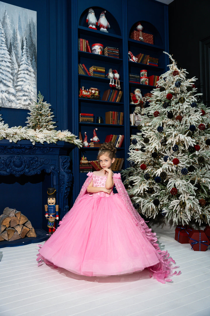 Child in a pink dress standing in a festively decorated room with a Christmas tree and bookshelf.