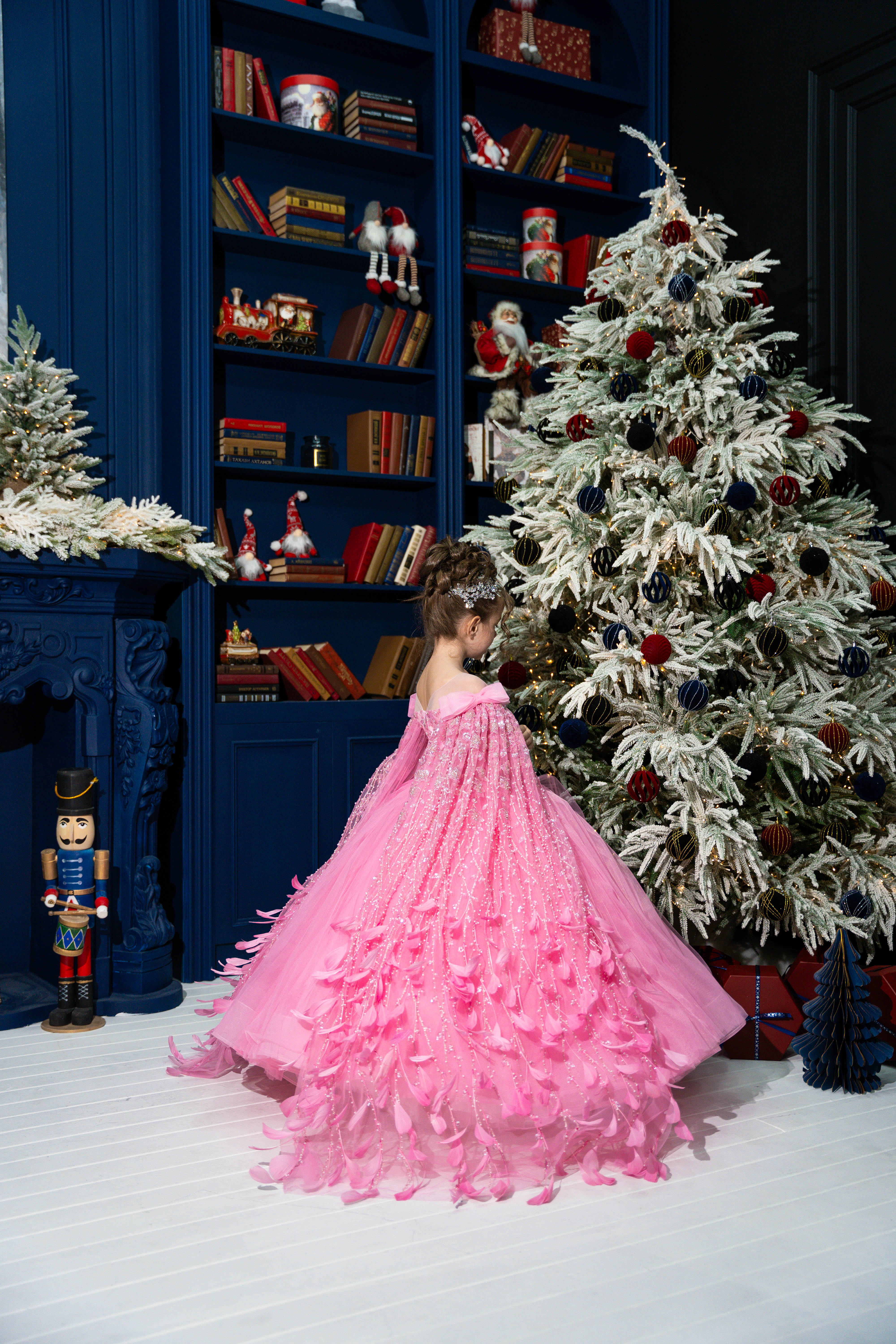 Child in a pink dress standing in front of a decorated Christmas tree and bookshelf.