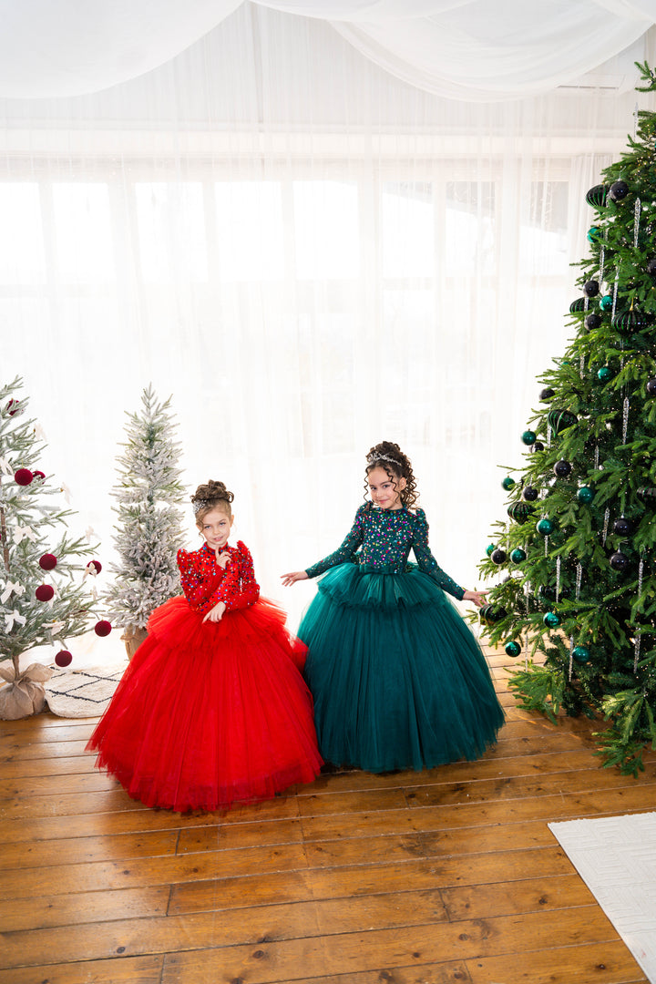 Two children in red and green dresses standing in front of Christmas trees.