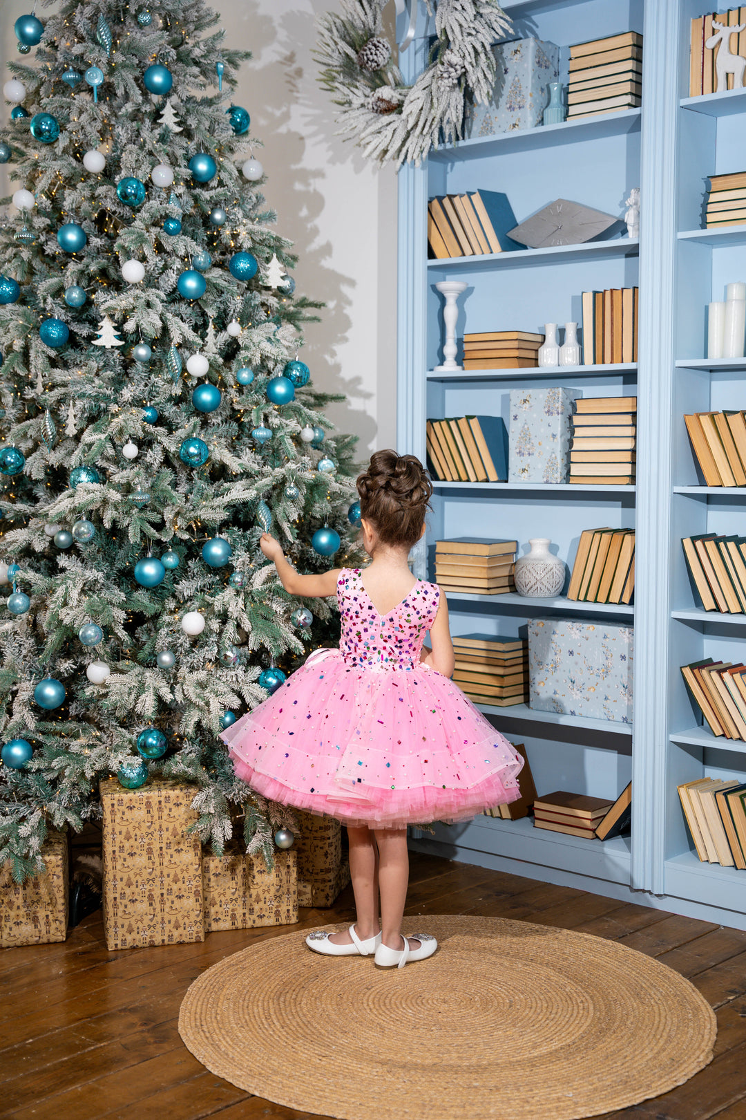 Child in a pink dress standing next to a decorated Christmas tree in a room with a bookshelf.
