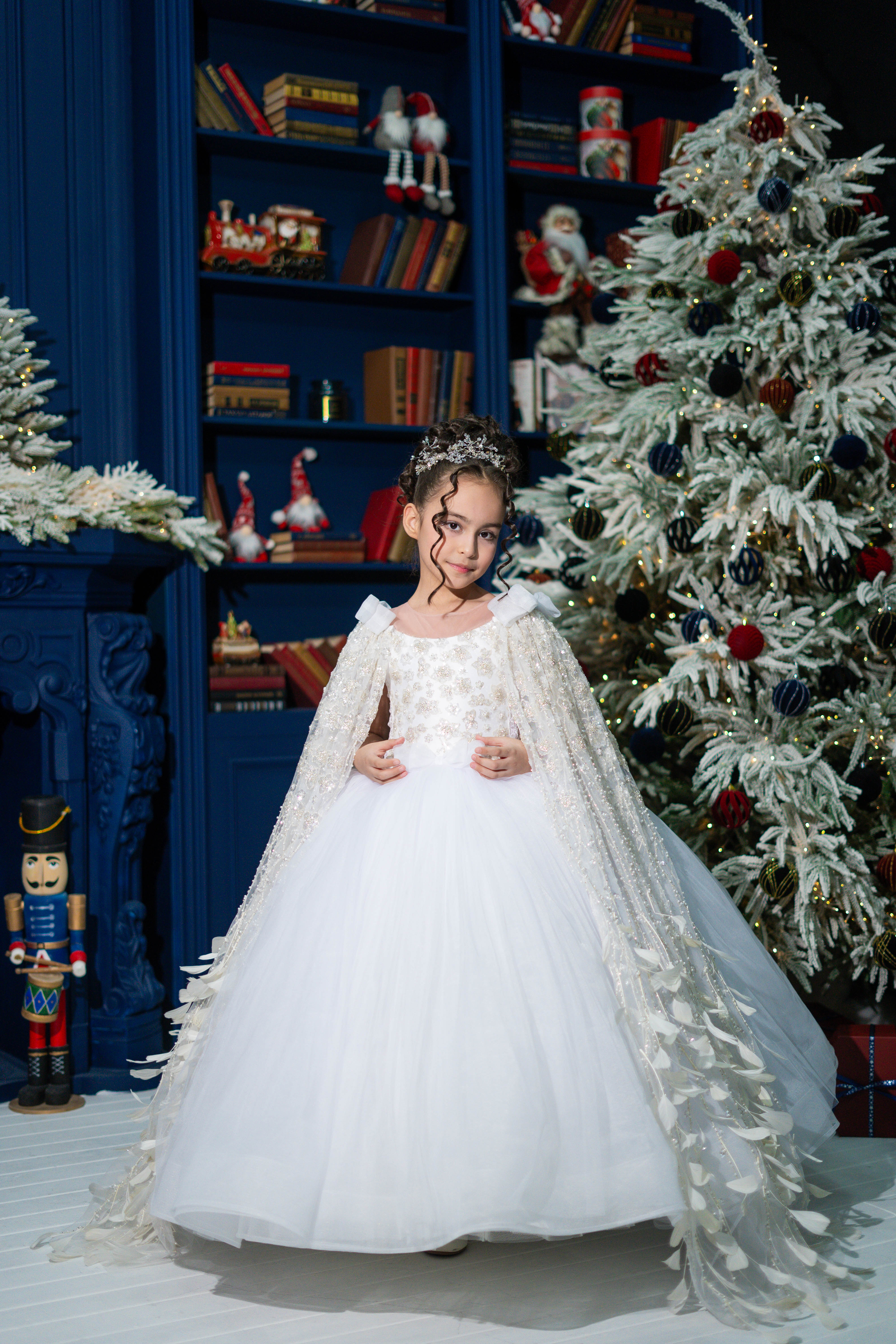 Child in a white dress standing in front of a decorated Christmas tree and bookshelf.