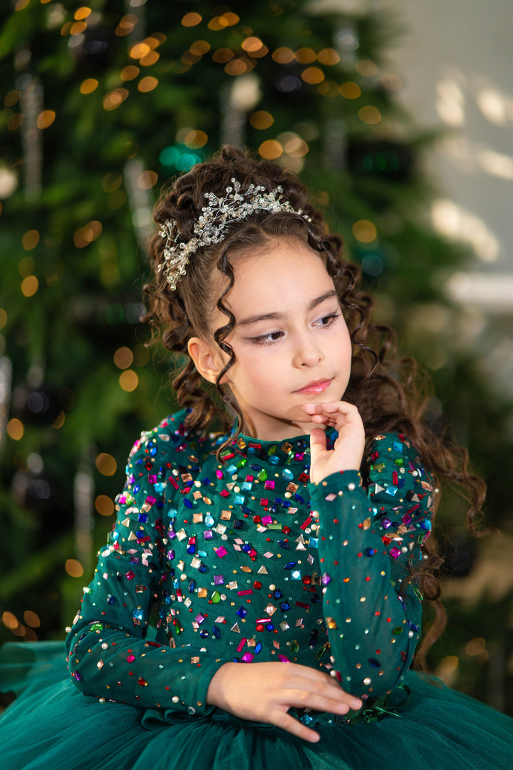 Young girl in a green dress with colorful sequins in front of a decorated Christmas tree.