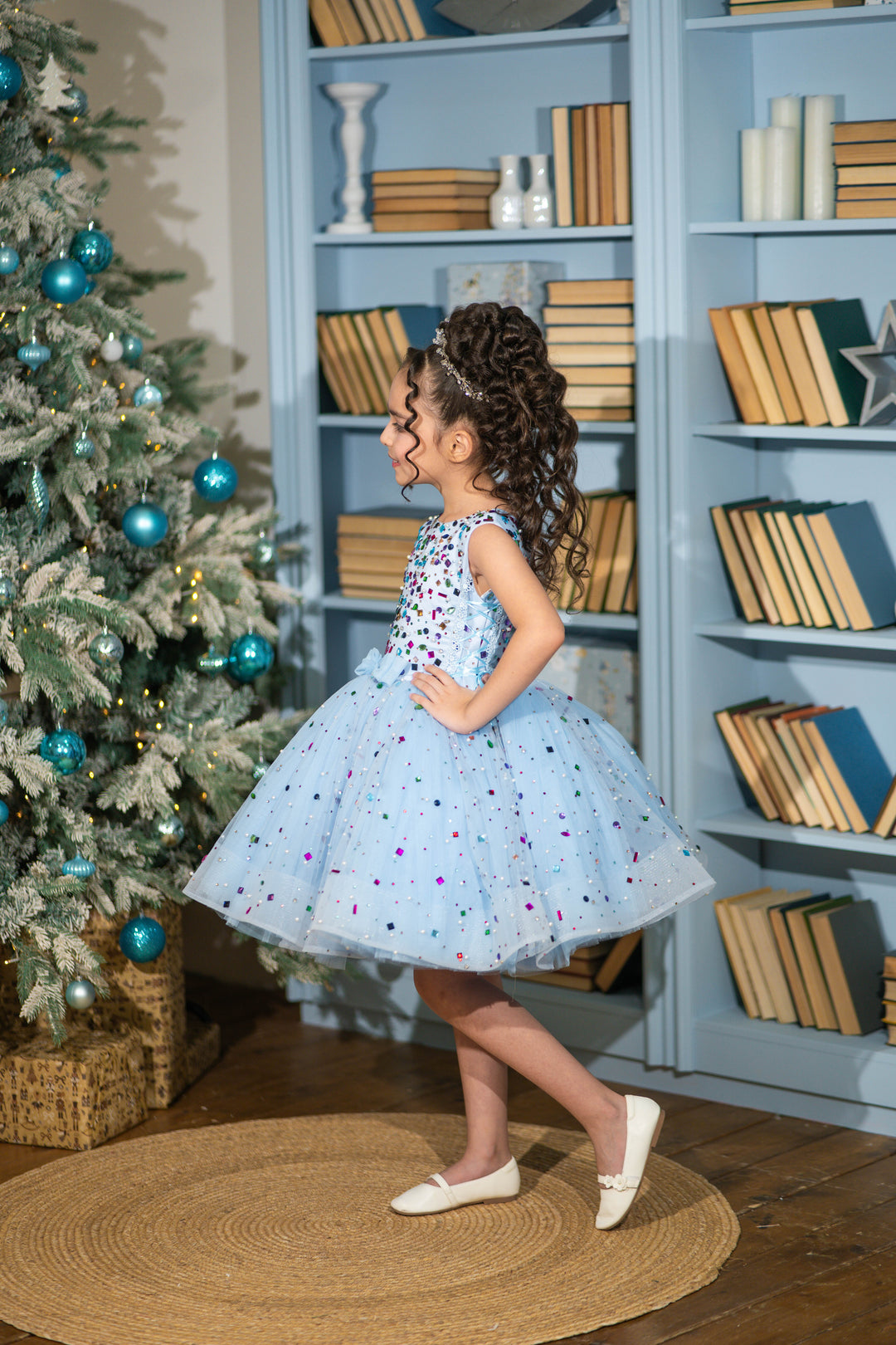 Child in a light blue dress with colorful polka dots standing in front of a bookshelf decorated with Christmas trees.