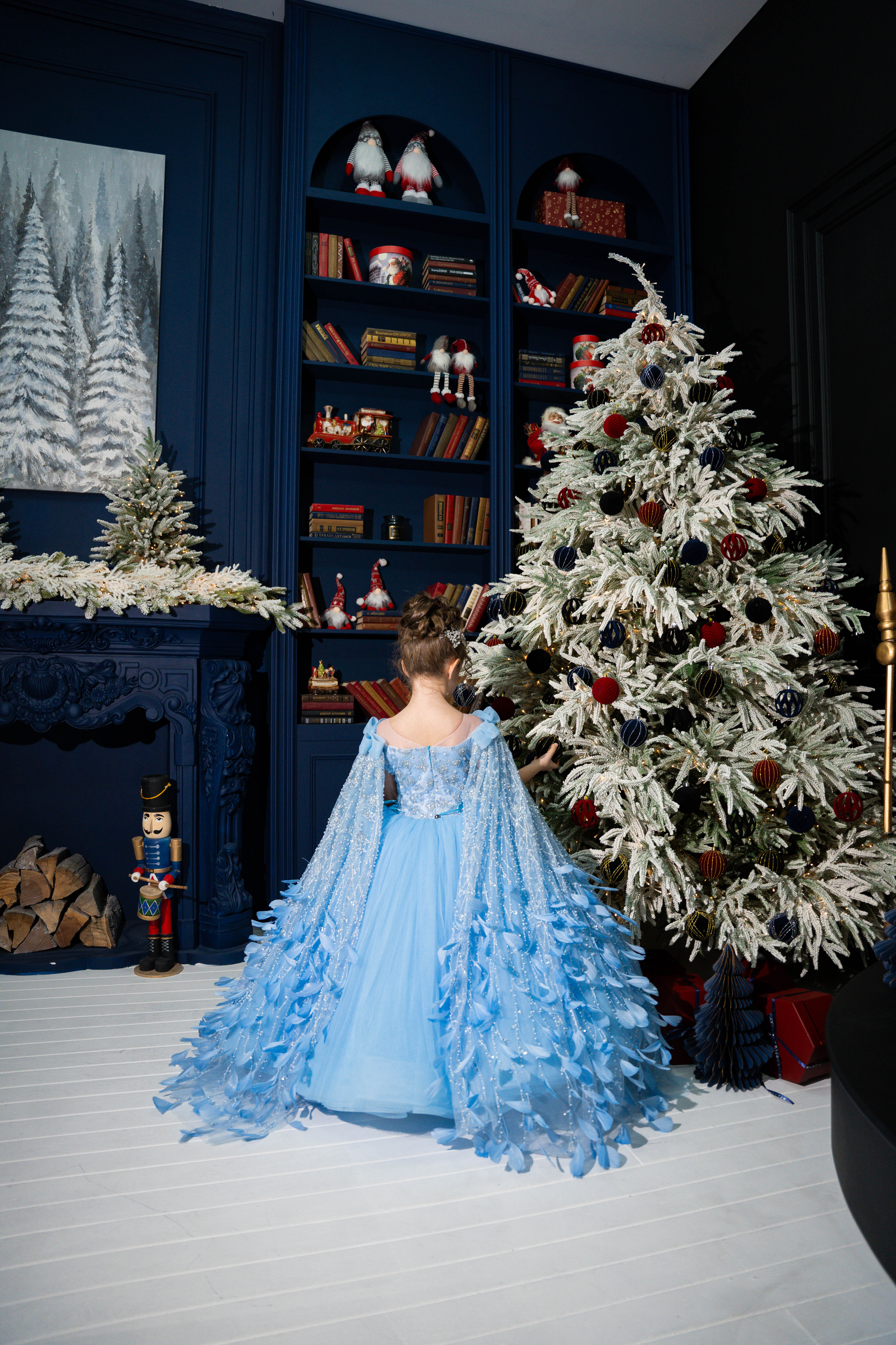 A girl in a blue dress standing in a room with a Christmas tree and decorated shelves.