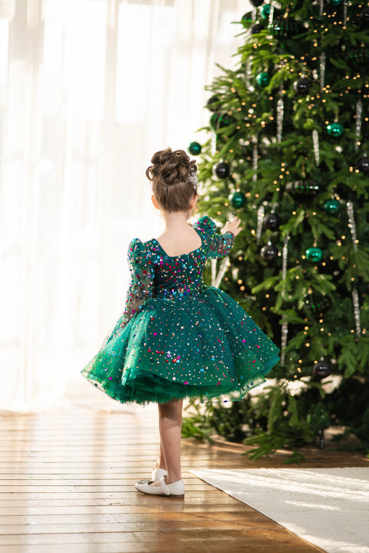 Child in a green dress standing in front of a decorated Christmas tree.