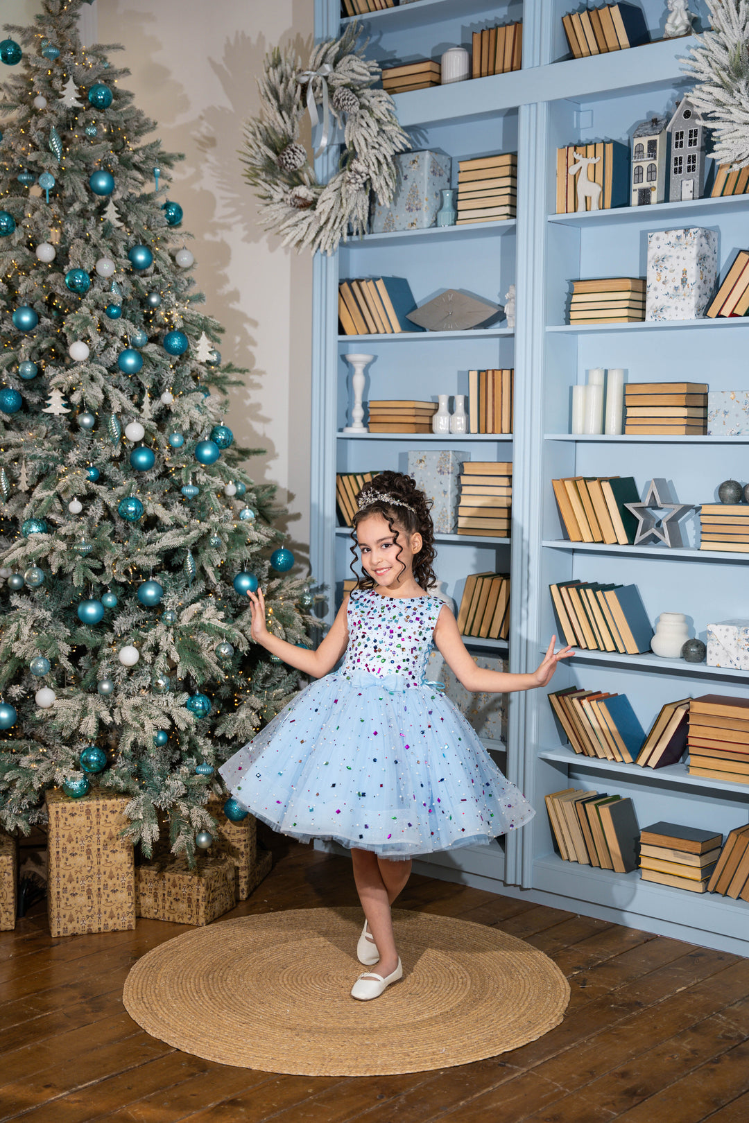 Young girl in a blue dress standing in front of a Christmas tree and bookshelf.