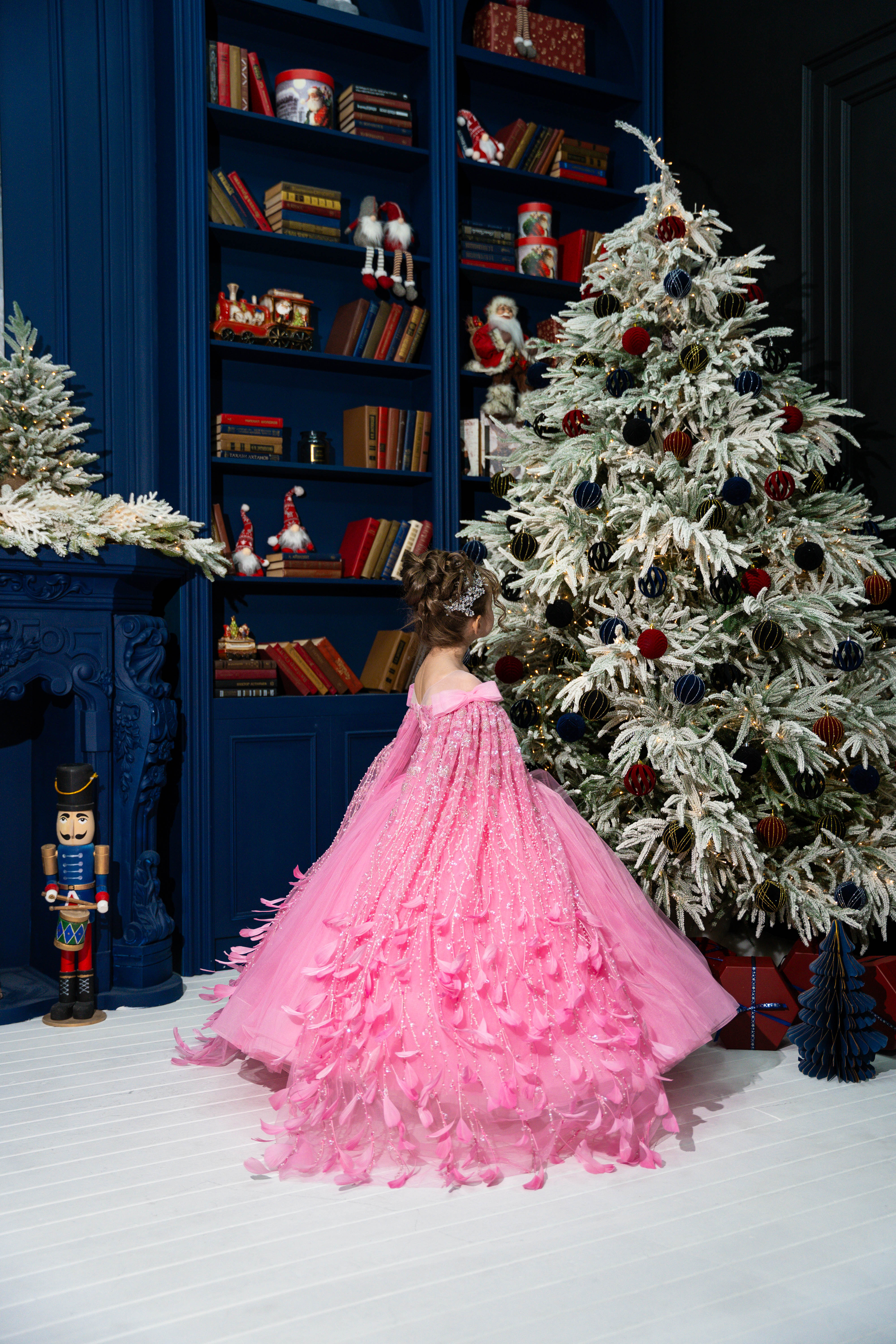Child in a pink dress standing in front of a decorated Christmas tree and bookshelf.