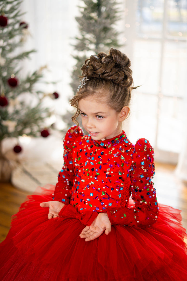 Young girl in a red dress with colorful embellishments sitting in front of a decorated Christmas tree.