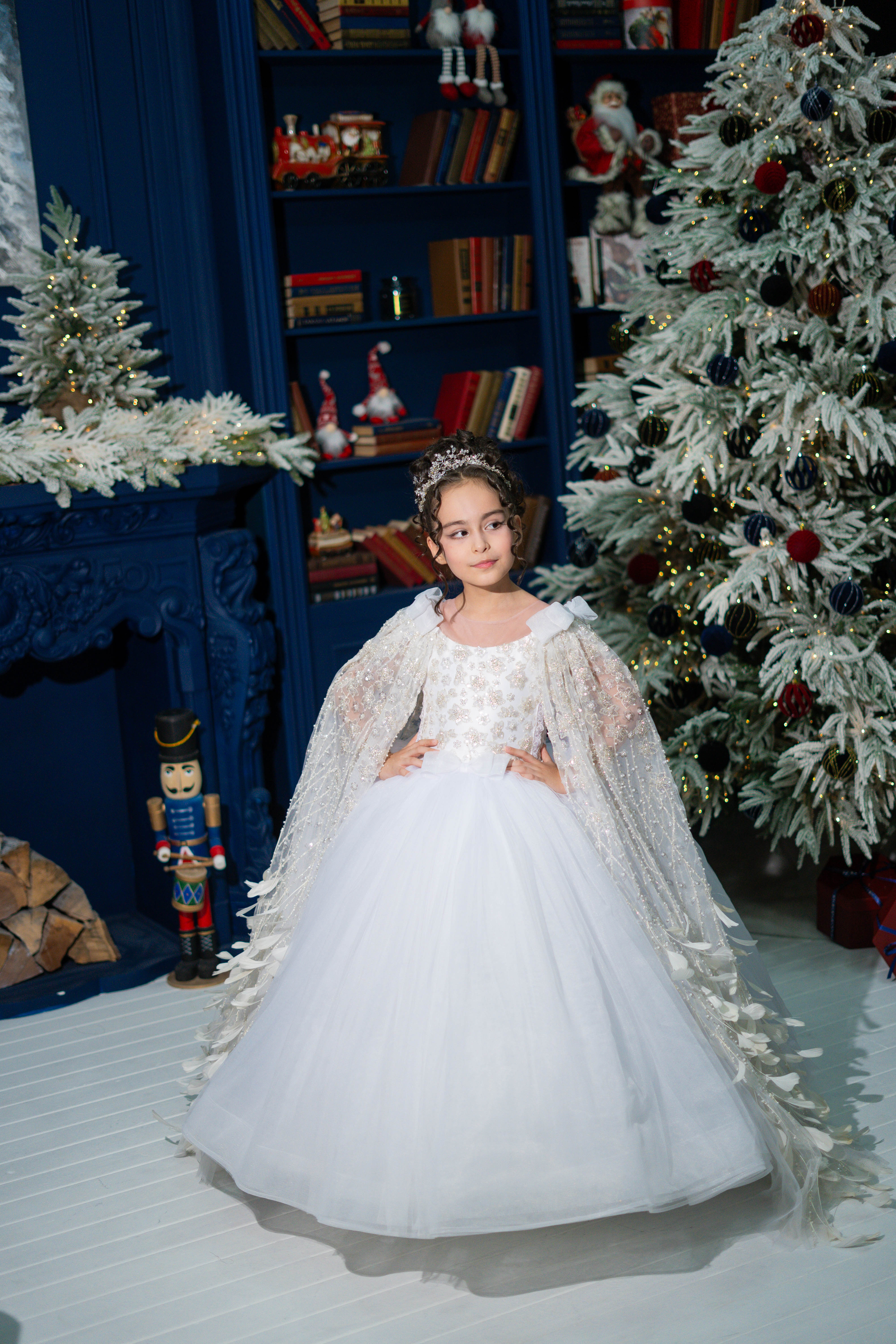 Young girl in a white dress standing in front of a decorated Christmas tree and bookshelf.
