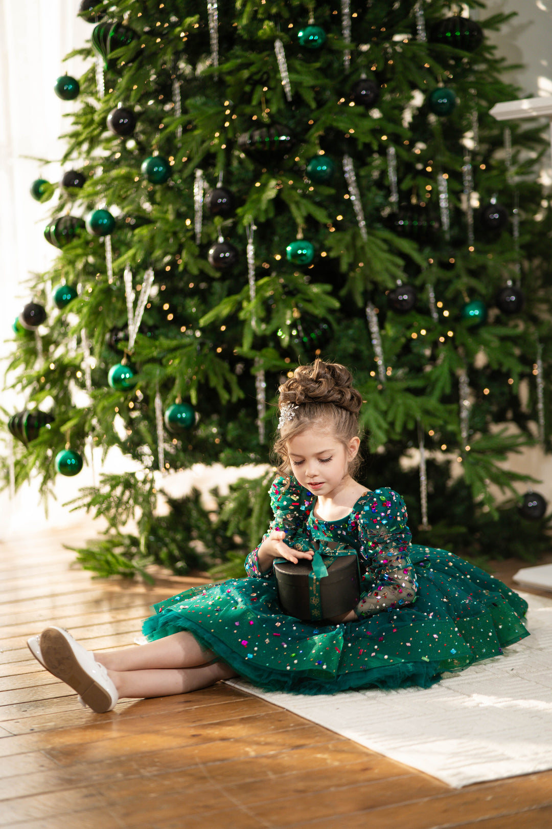 Young girl in a green dress sitting on the floor in front of a decorated Christmas tree.
