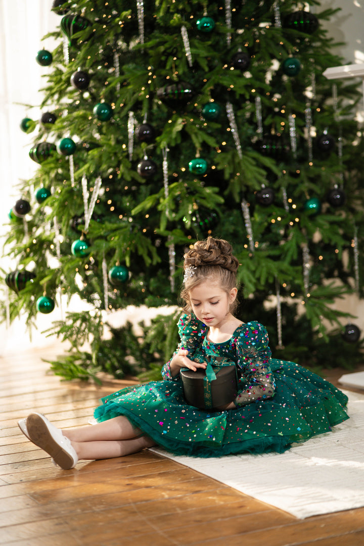 Young girl in a green dress sitting on the floor in front of a decorated Christmas tree.