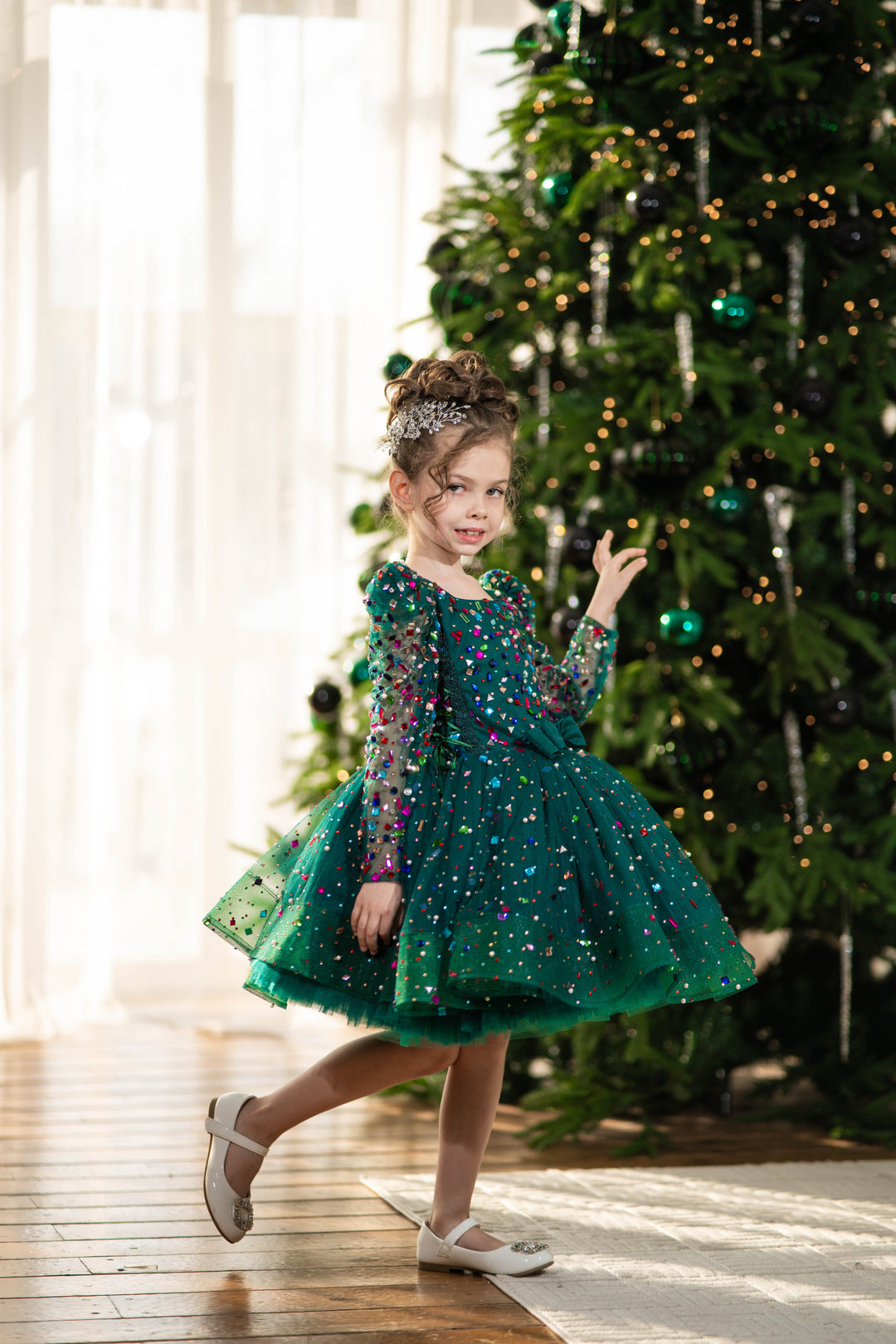 Baby girl in a green dress standing in front of a decorated Christmas tree.