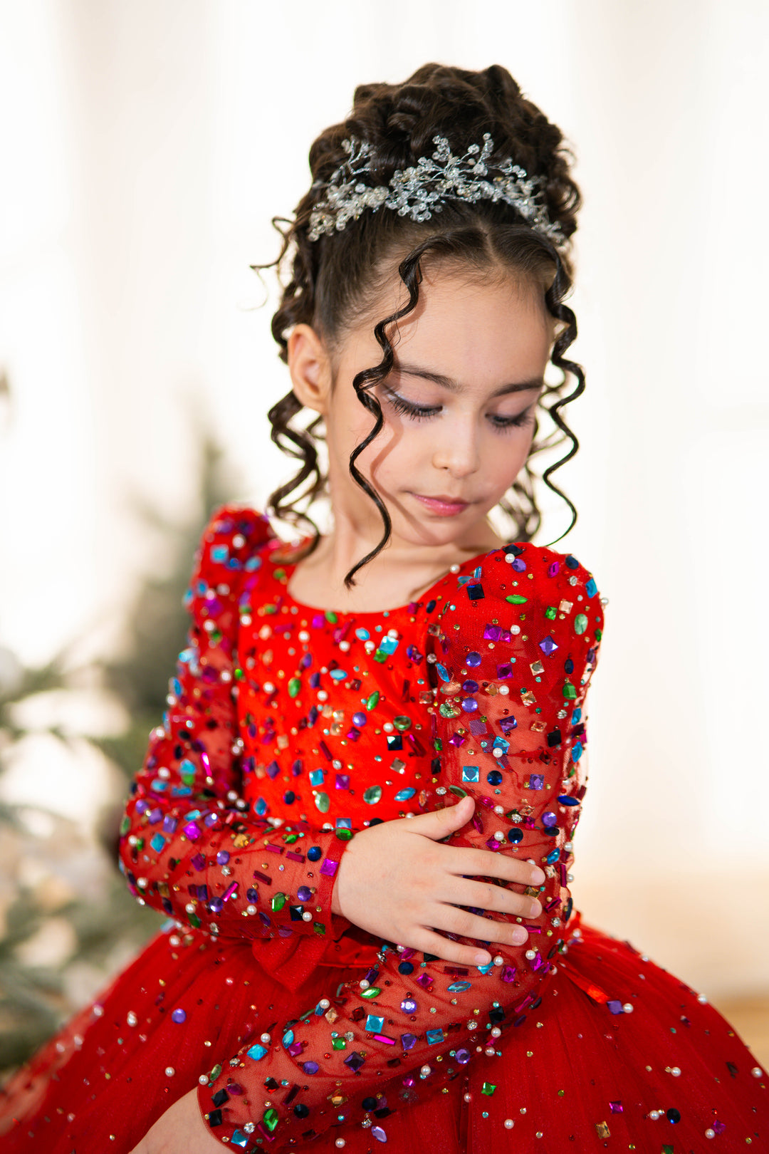 Young girl in a red dress with colorful sequins, wearing a tiara, against a blurred background.