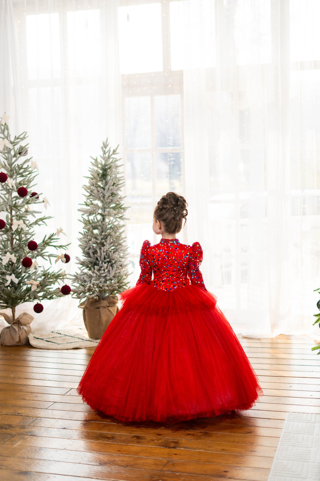 Young girl in a red dress standing in front of Christmas trees with white curtains.
