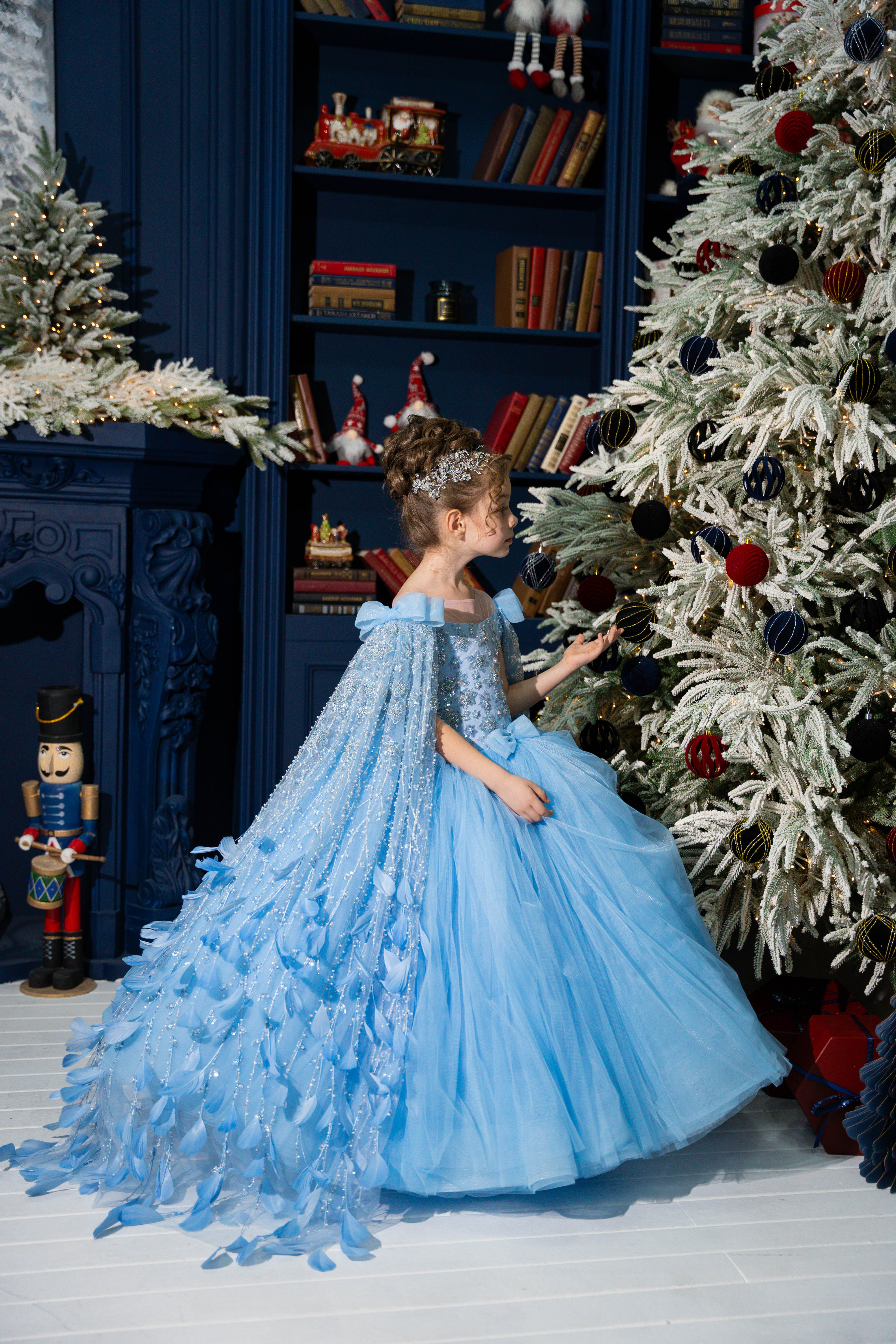 Young girl in a blue dress standing in front of a decorated Christmas tree.