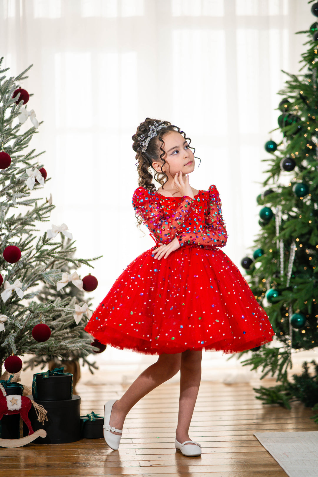 Baby girl in a red dress standing between two decorated Christmas trees.