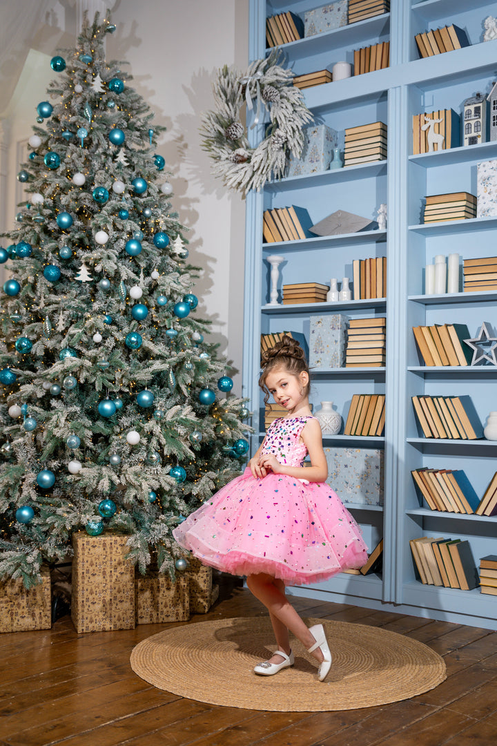 Baby girl in a pink dress standing in front of a decorated Christmas tree and bookshelf.