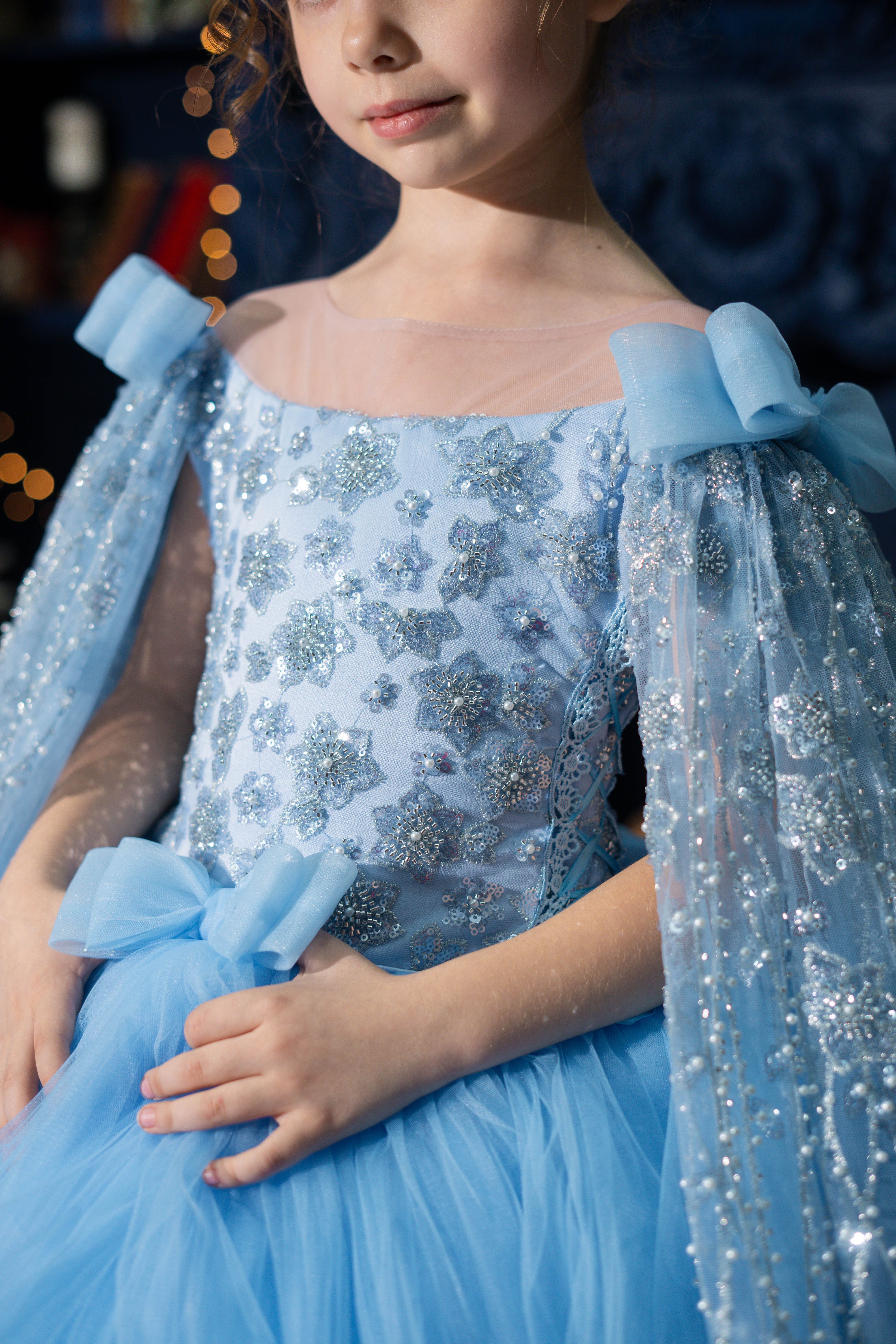 Child wearing a blue sequined dress with cape-like sleeves against a dark background.