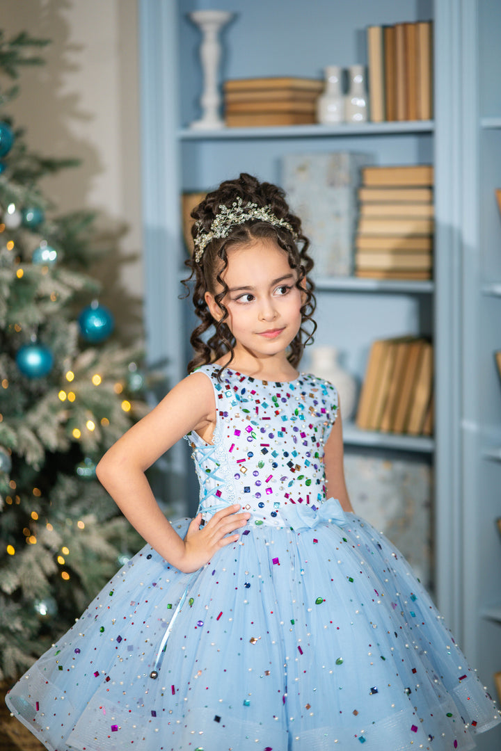 Young girl in a colorful dress standing in front of a decorated Christmas tree and bookshelf.