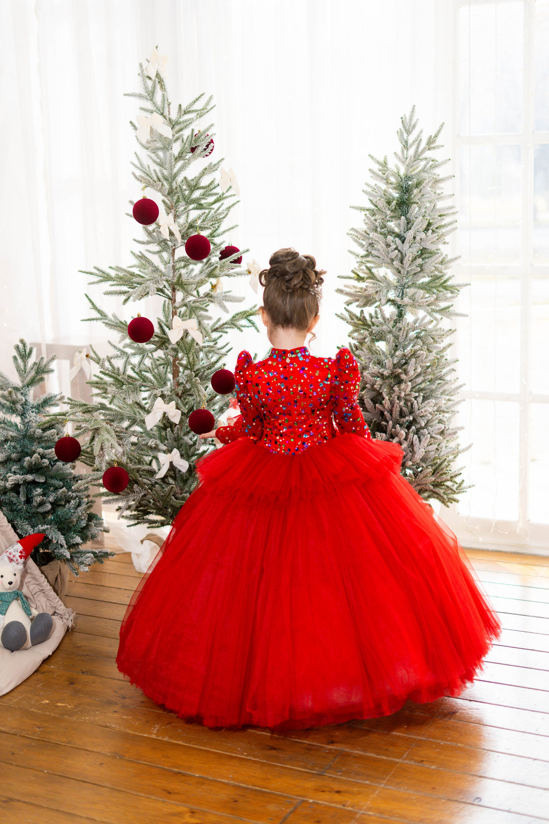 Child in a red dress standing in front of Christmas trees.