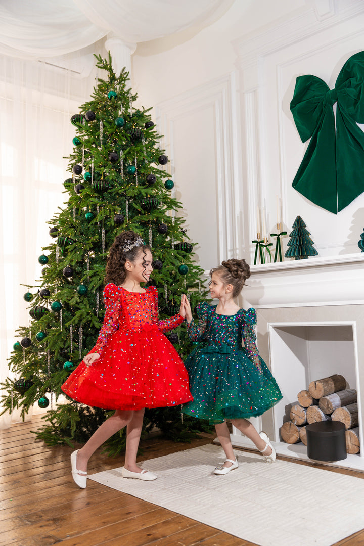Two children in red and green dresses standing in front of a decorated Christmas tree.