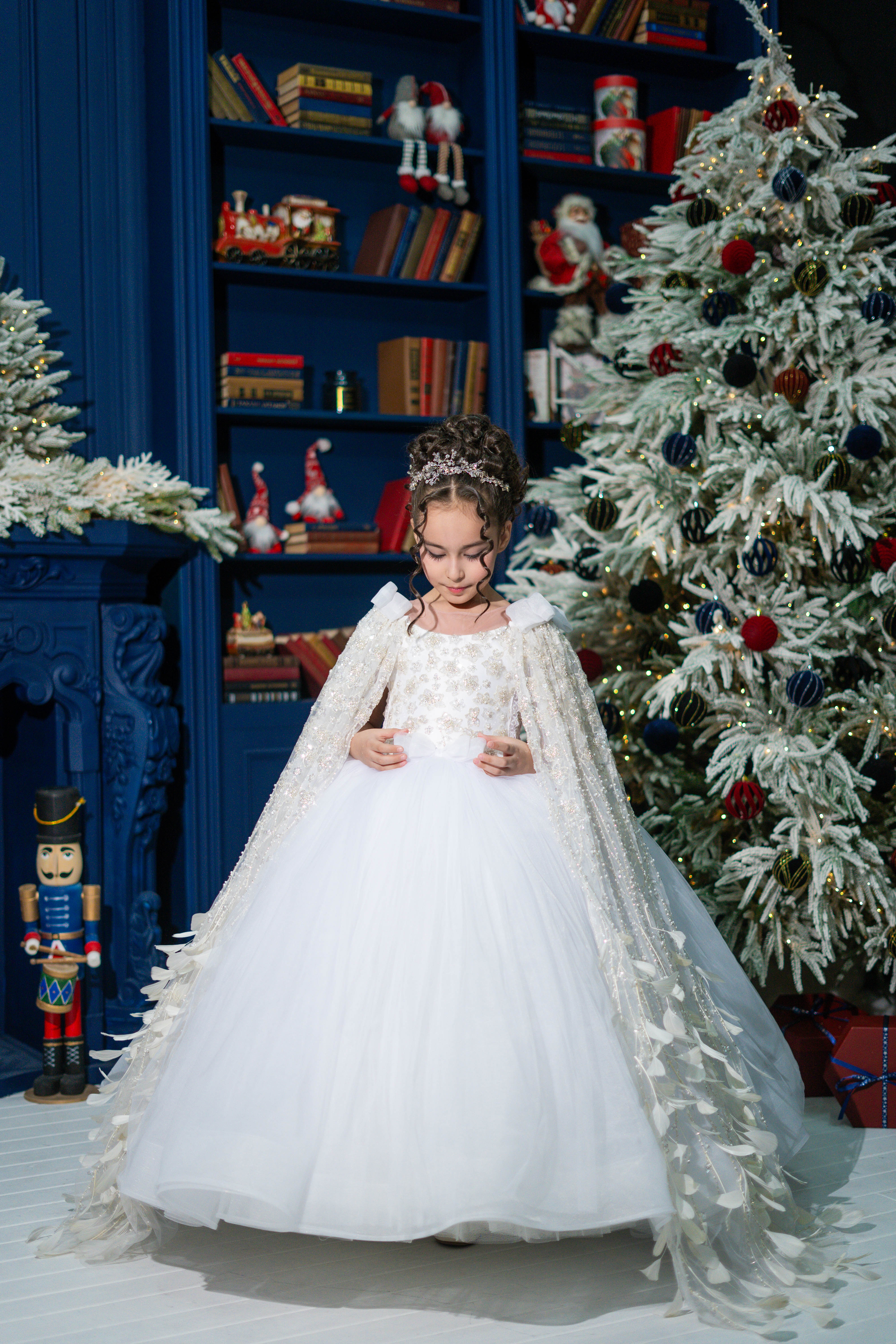 Young girl in a white dress with a cape standing in front of a decorated Christmas tree and bookshelf.