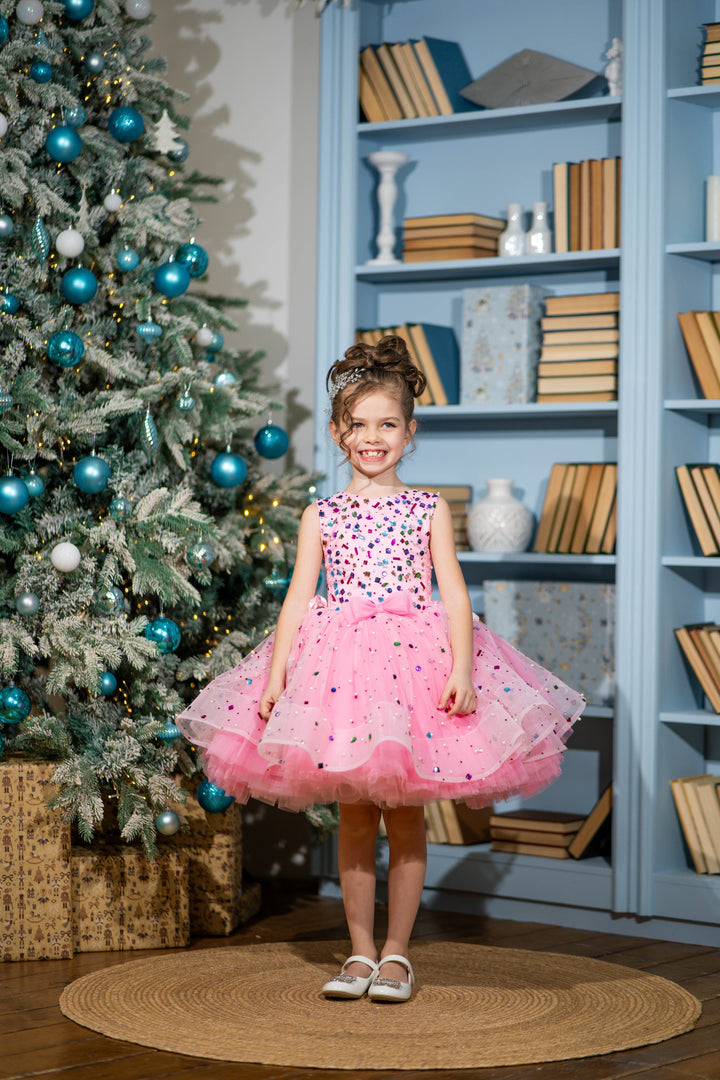 Young girl in a pink dress standing in front of a decorated Christmas tree and bookshelf.
