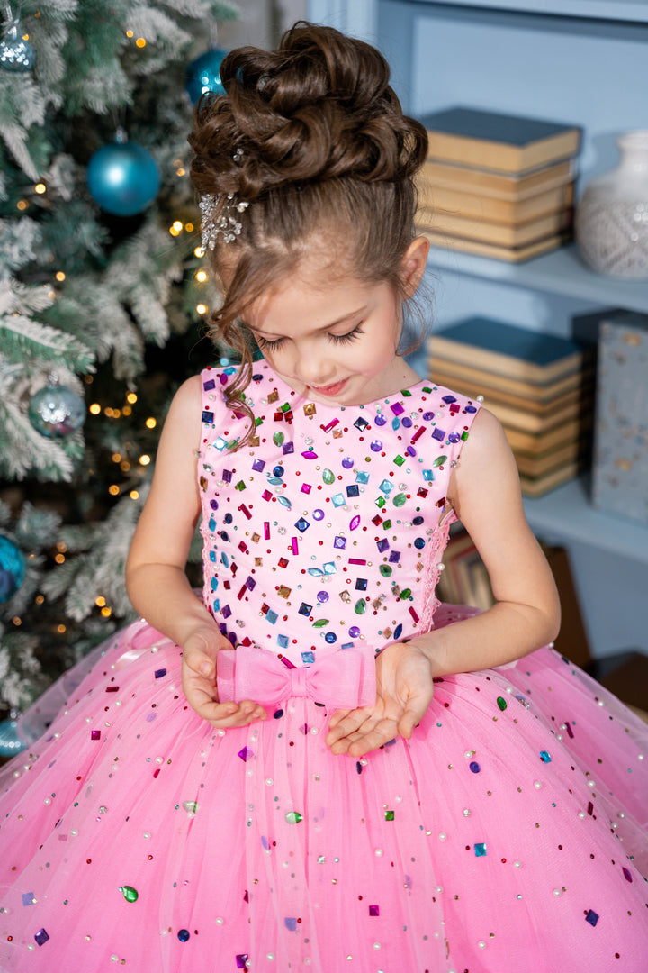 Baby girl in a pink dress with colorful sequins standing in front of a decorated Christmas tree.