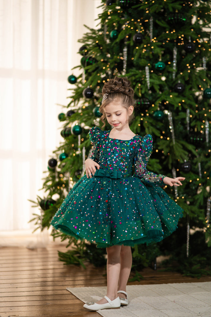 Baby girl in a green dress standing in front of a decorated Christmas tree.