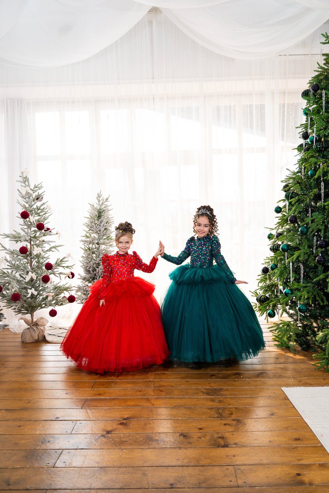 Two children in red and green dresses standing in front of Christmas trees.