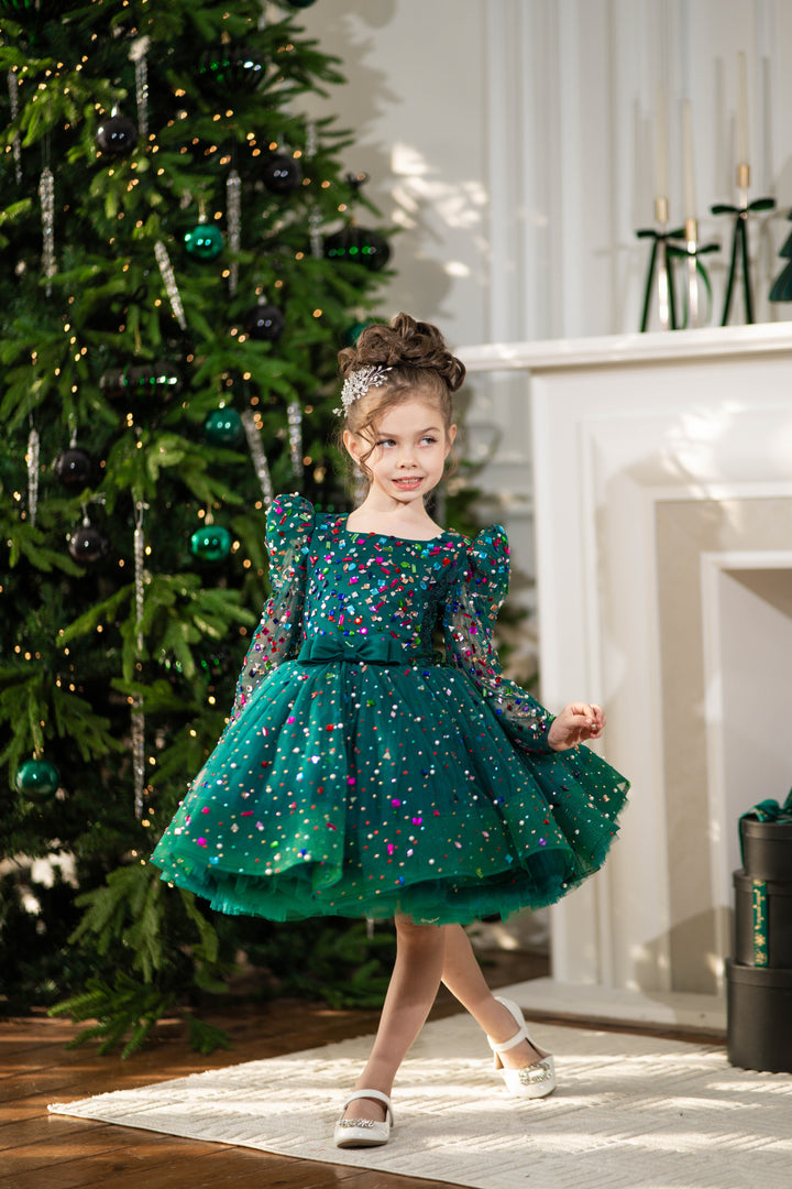 Baby girl in a green sequined dress standing in front of a decorated Christmas tree.