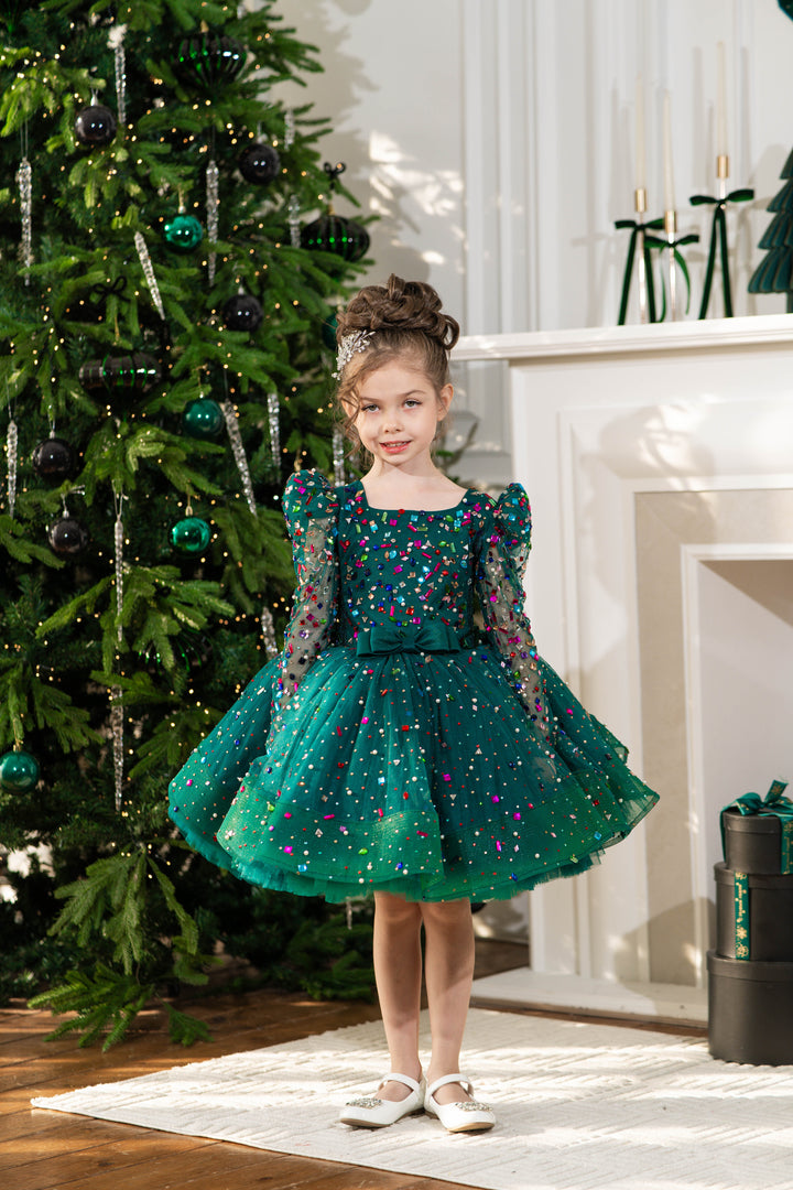 Baby girl in a green sequined dress standing in front of a decorated Christmas tree.