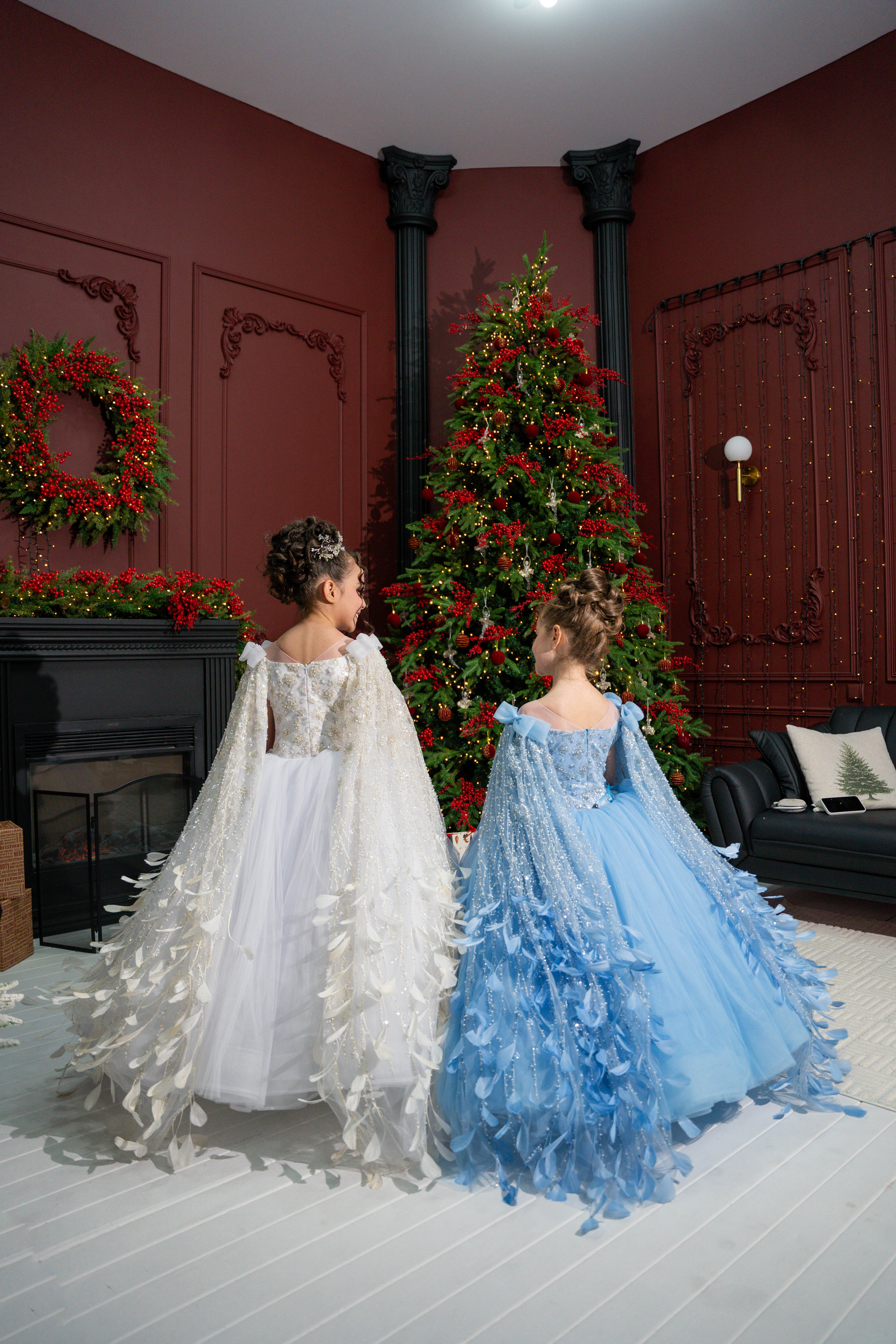 Two children in white and blue dresses standing in a room decorated for Christmas.