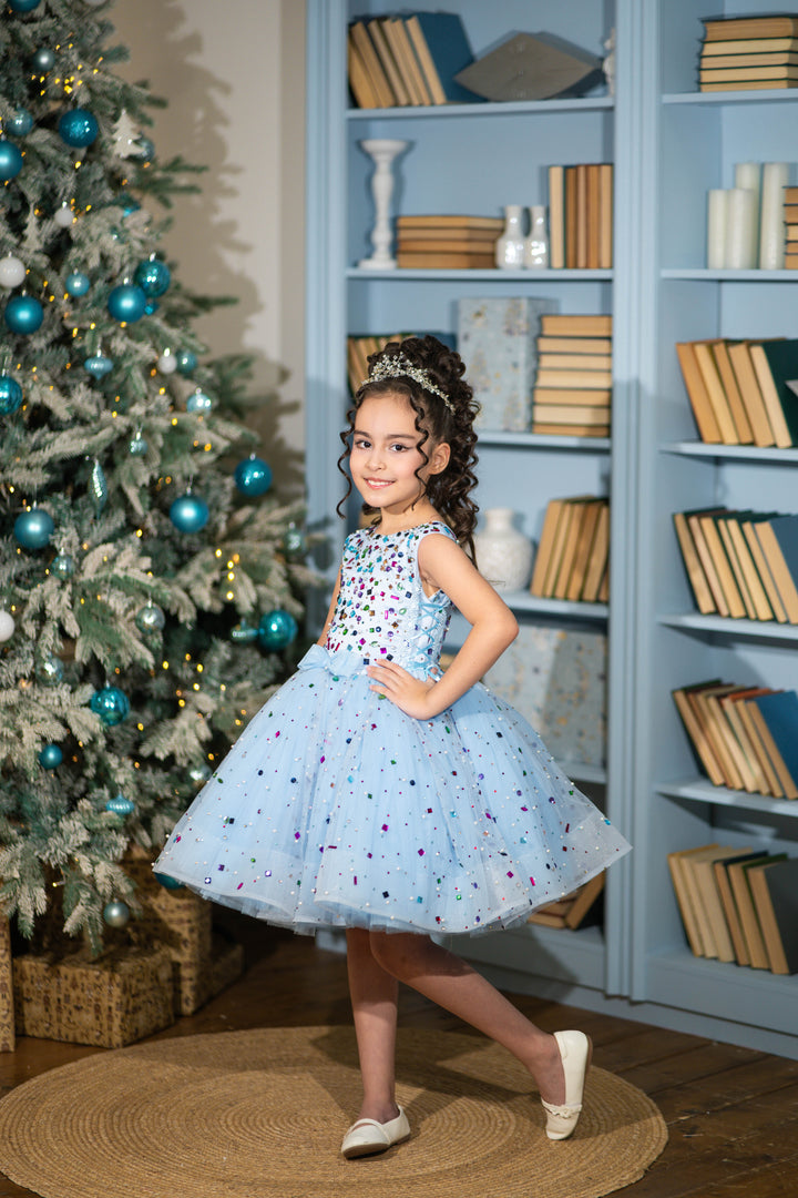 Young girl in a polka dot dress standing in front of a decorated Christmas tree and bookshelf.