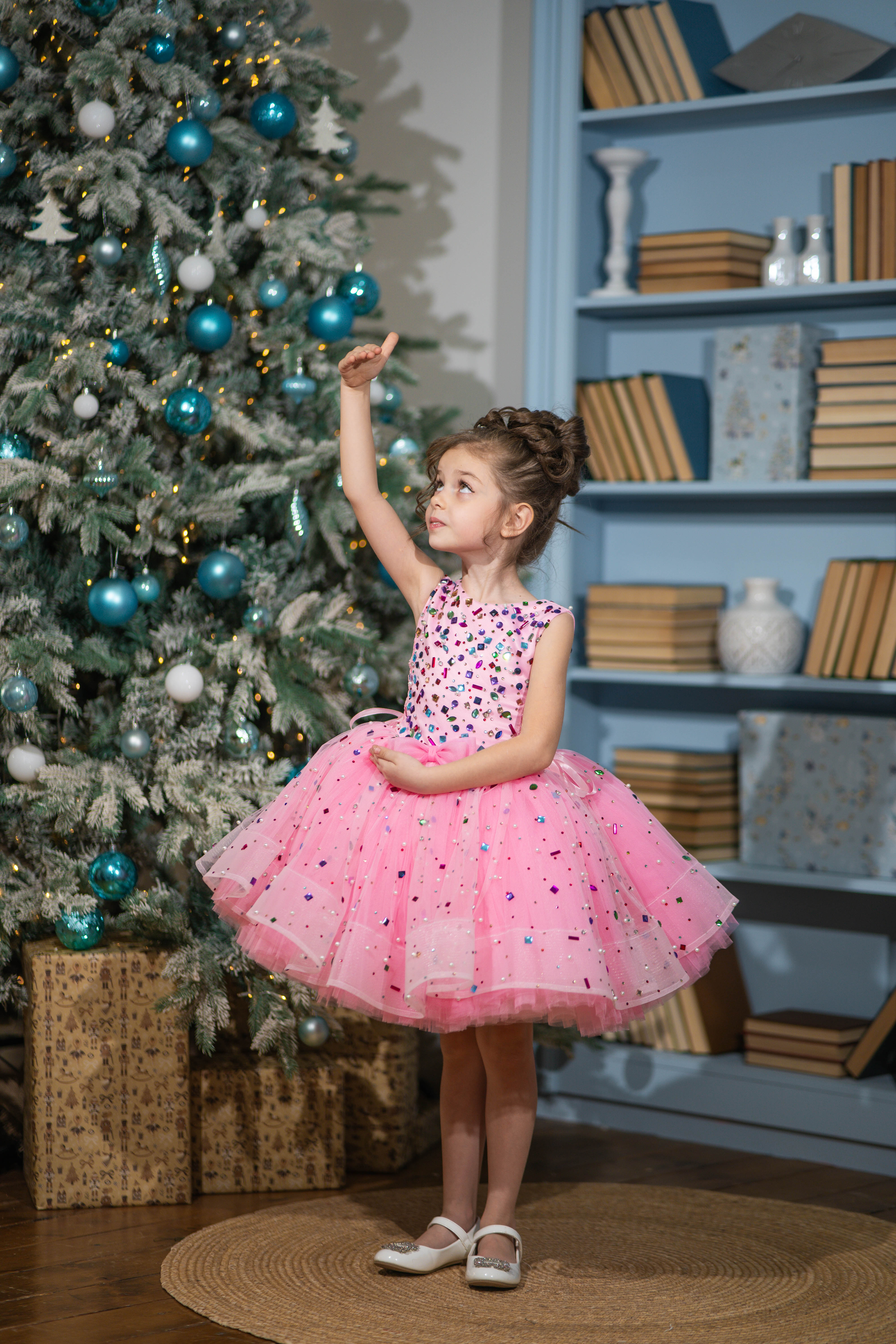 Baby girl in a pink dress standing next to a decorated Christmas tree.