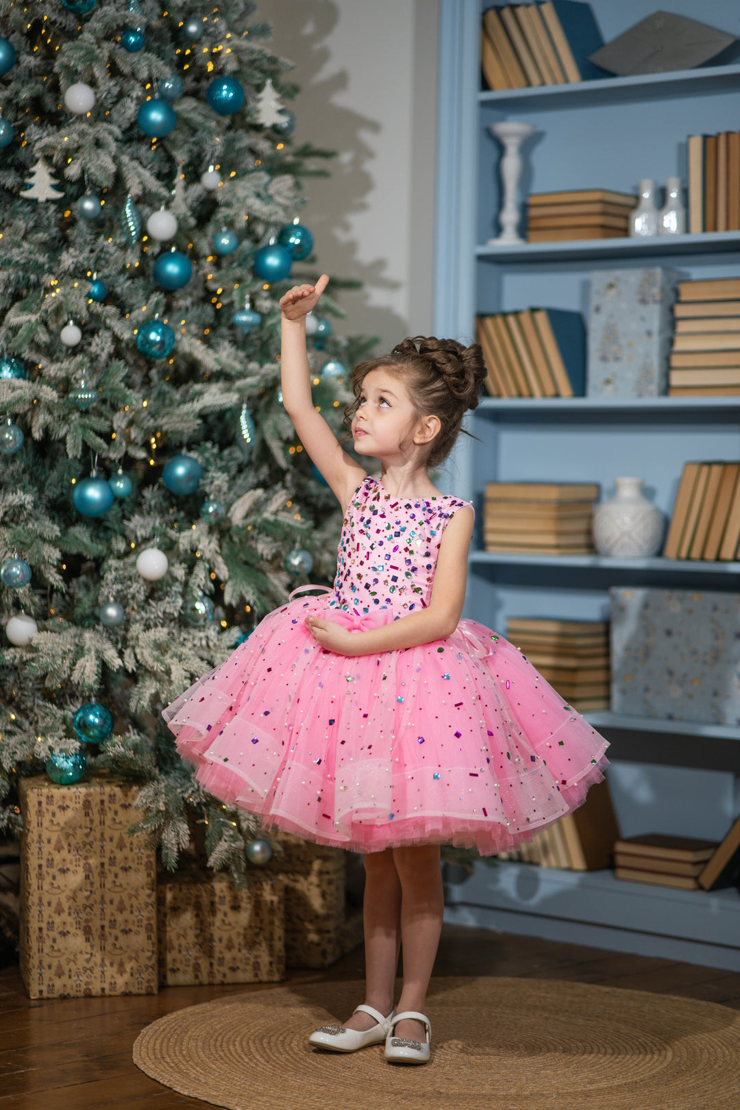 Baby girl in a pink dress standing next to a decorated Christmas tree.